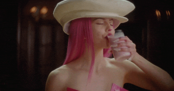Woman with pink hair wearing a wide-brimmed hat, drinking a glass of water, in a dimly lit setting.