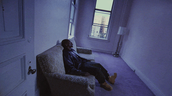 Person sitting on a patterned couch in an empty room with white walls and window, looking outside.