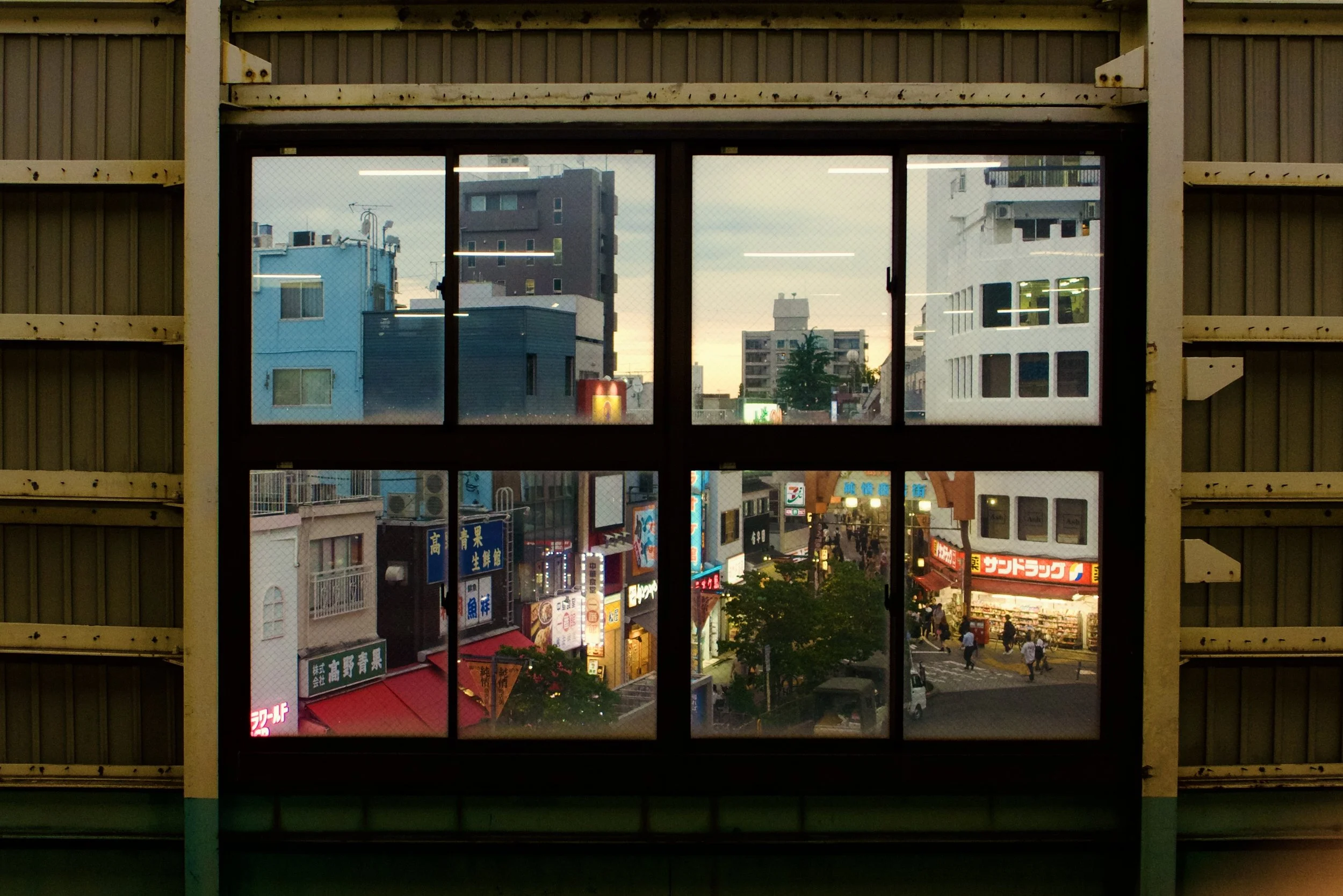 View of a city street through a window with a wire mesh, showing colorful buildings, neon signs, and pedestrians in an urban area at dusk.
