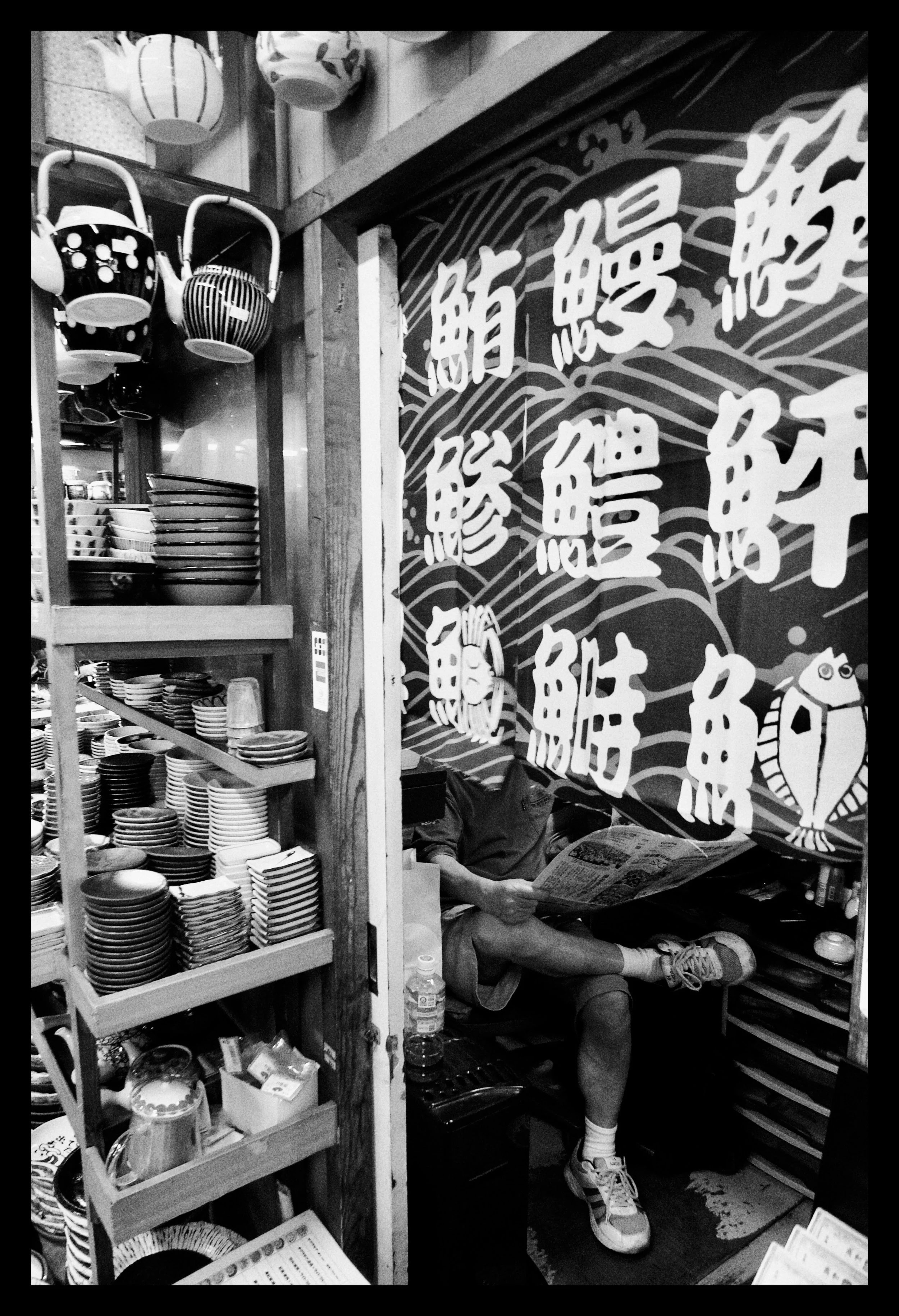 Person sitting in a small, cluttered space behind a counter, reading a newspaper, surrounded by dishes and kitchenware, with Japanese lanterns hanging above.