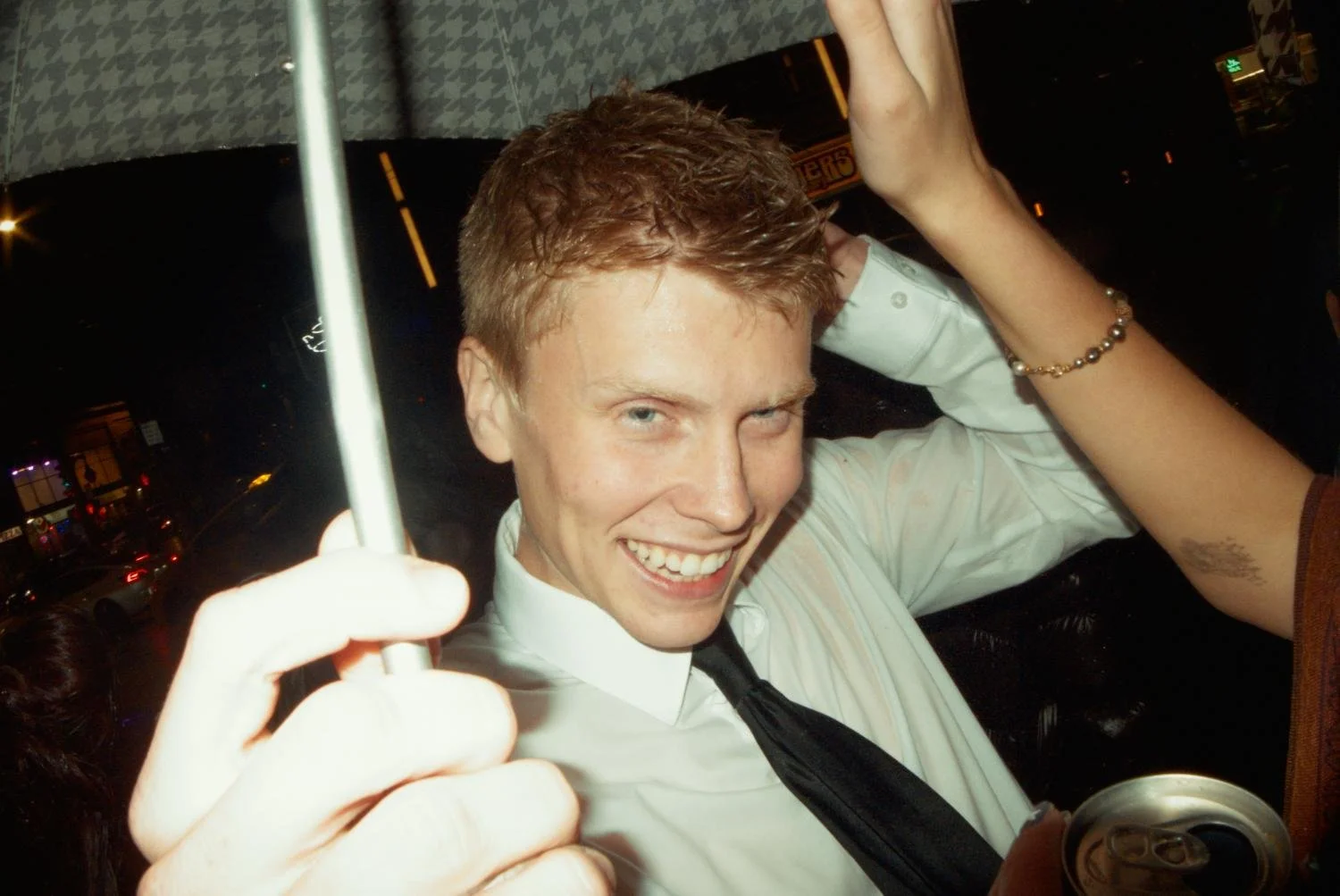A young man with red hair smiling at the camera while holding an umbrella, with another person's arm and hand nearby, at night with city lights in the background.