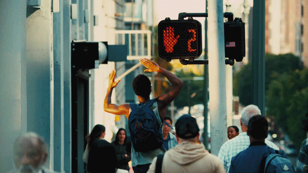 A young man with a backpack crossing a busy city street, raising his hand to stop traffic, while pedestrians wait at the crosswalk.
