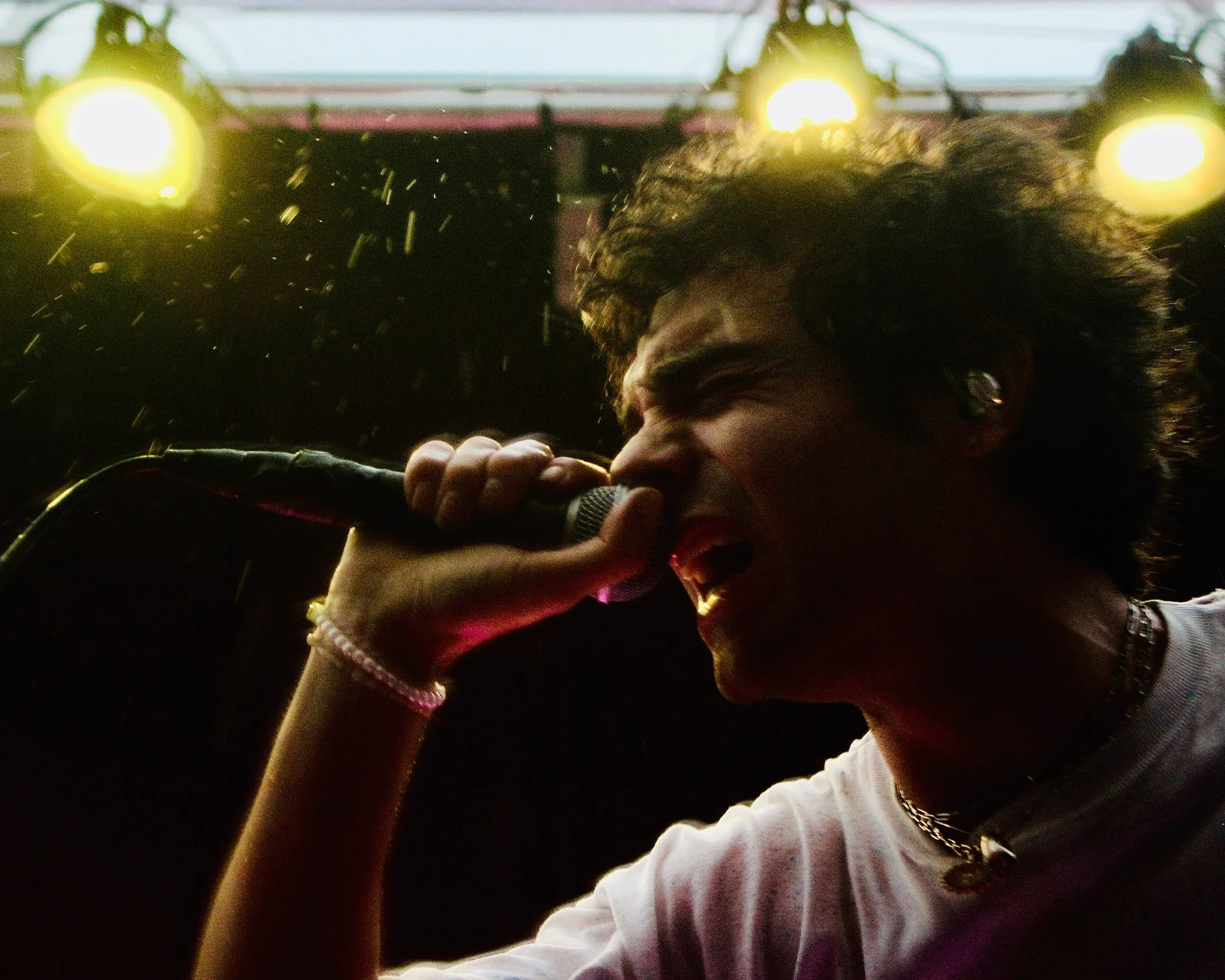 A person with curly hair, earrings, and a beaded bracelet, singing into a microphone while driving a vehicle, with bright yellow dashboard lights and a windshield showing clouds and sky outside.