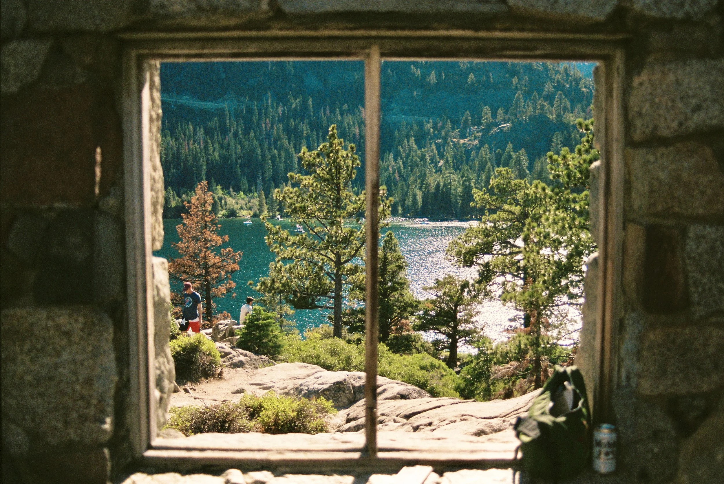 View of a lake and forest seen through a small stone window with a grid, overlooking a lakeside park with people and trees, surrounded by mountains.
