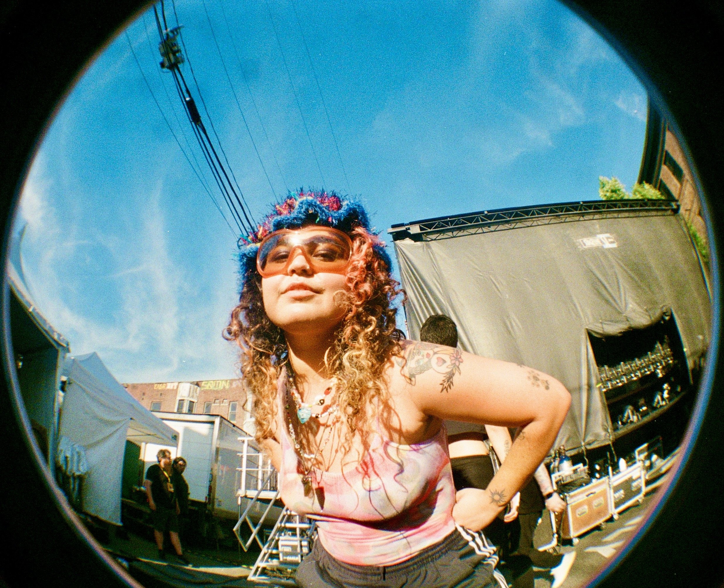 A person with curly hair and sunglasses taking a photo with a fisheye lens in an outdoor setting with people and buildings in the background.