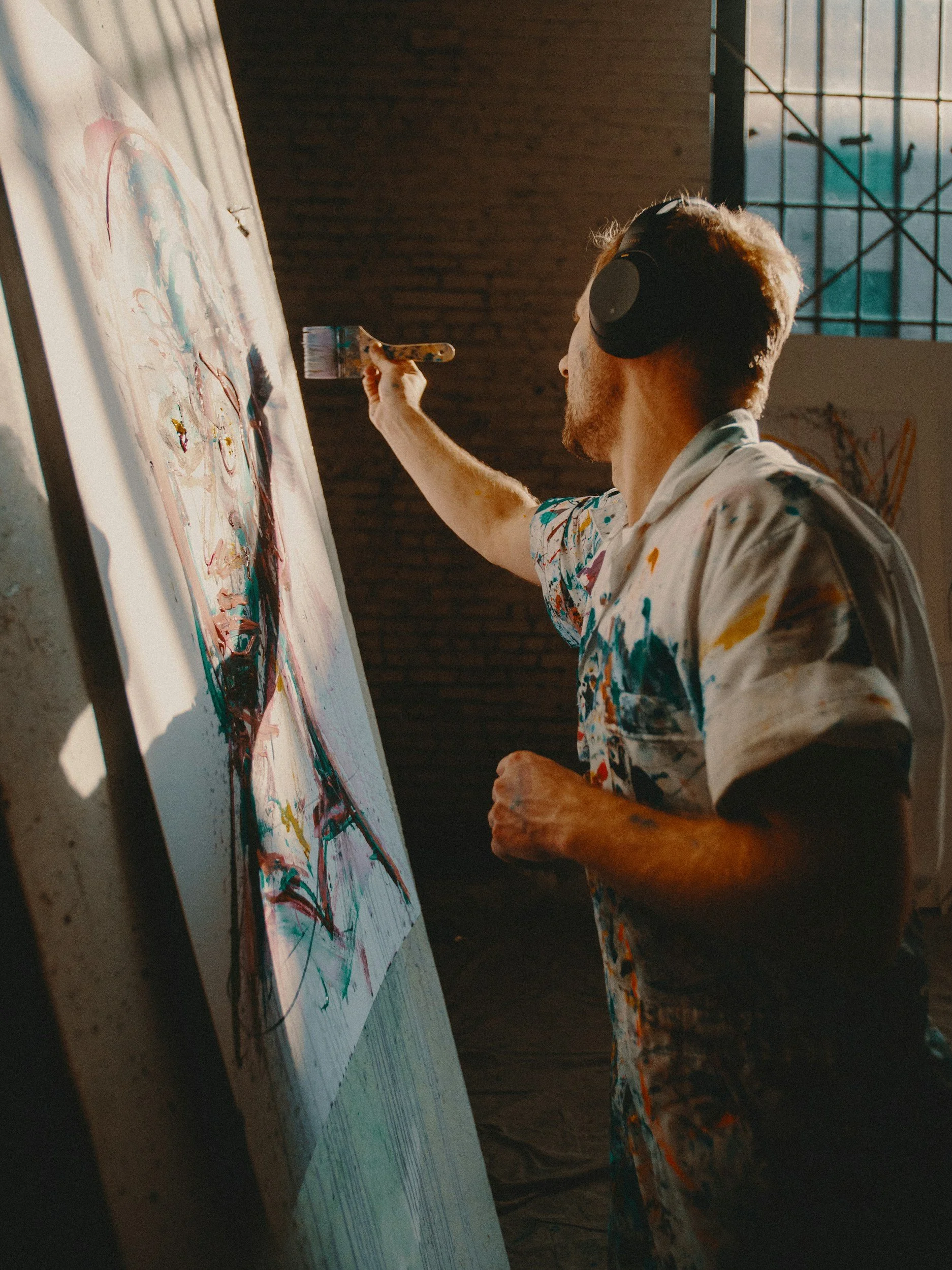 A male artist wearing a paint-splattered shirt and headphones is painting a colorful portrait on a large canvas in a studio with brick walls and large windows.