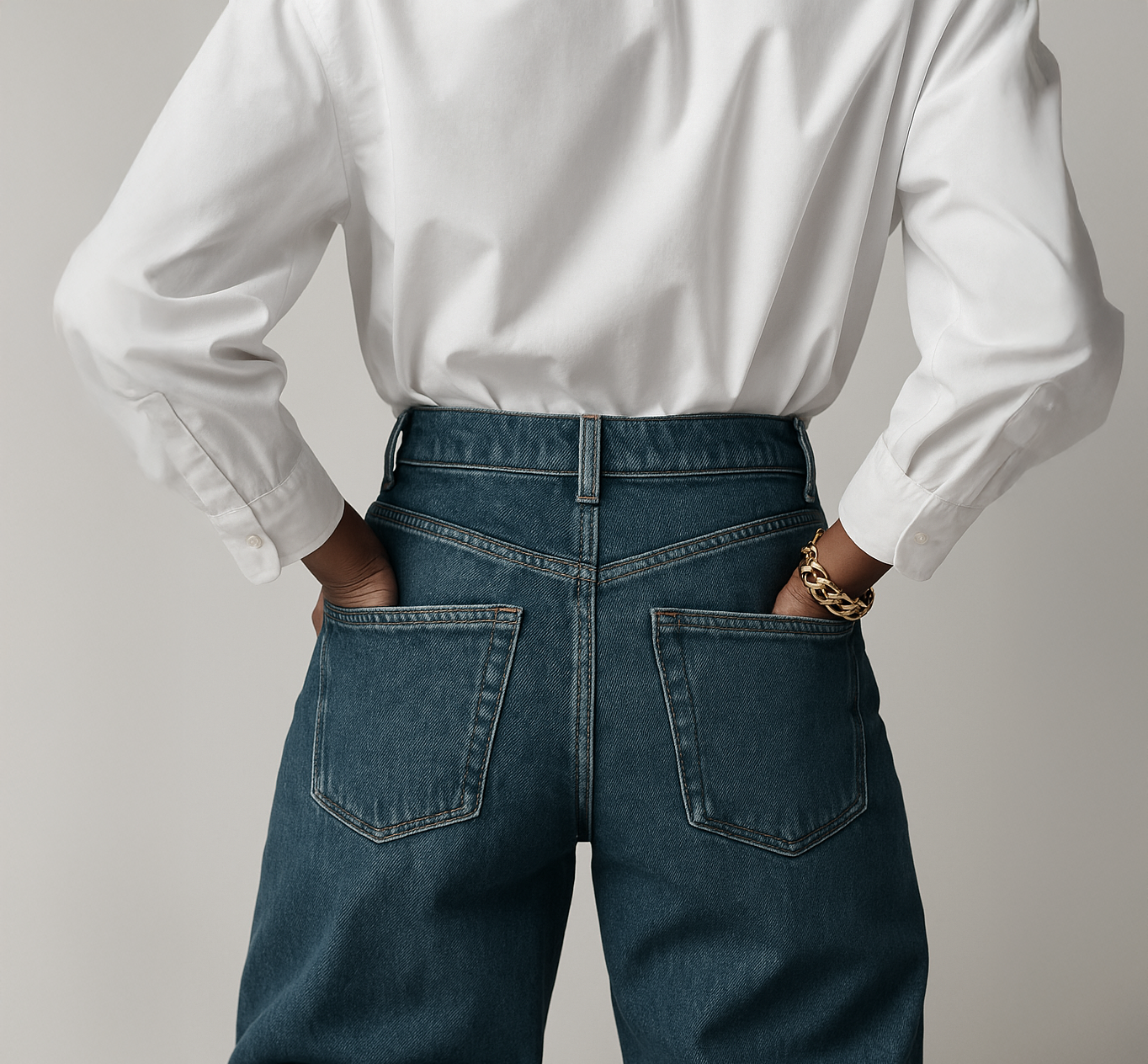 Person wearing a white shirt and high-waisted blue jeans with hands in back pockets, gold chain bracelet on right wrist.