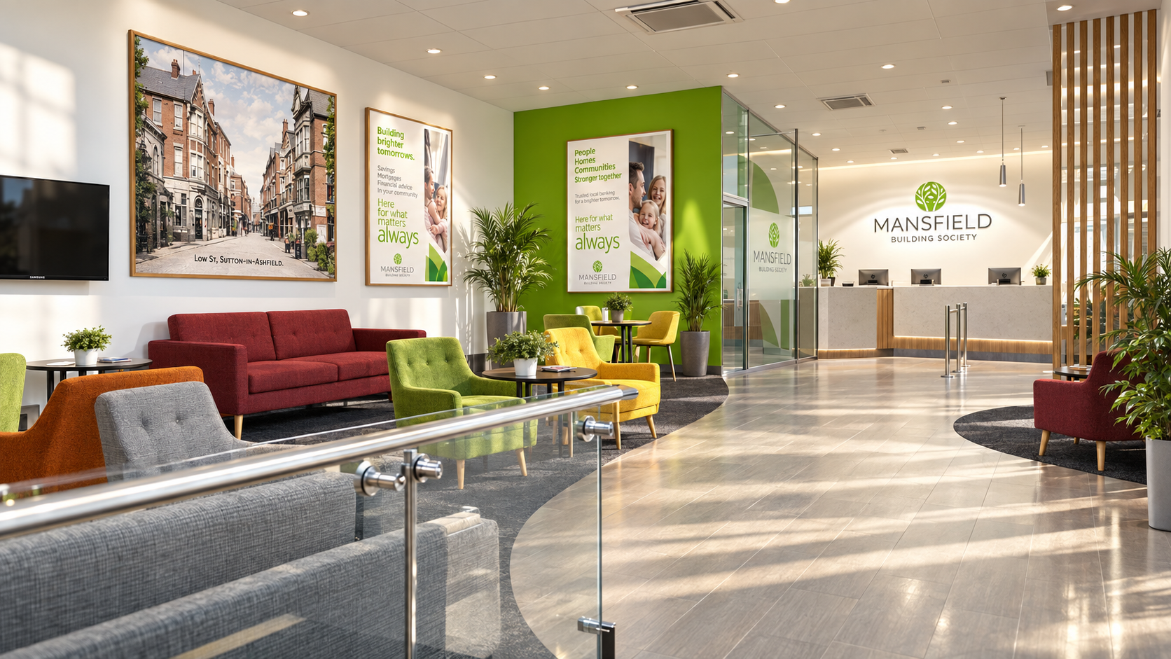Modern lobby of Mansfield Building Society featuring a seating area with colorful chairs and a red sofa, framed pictures and posters on the wall, indoor plants, and a reception desk with three monitors and the Mansfield Building Society logo.
