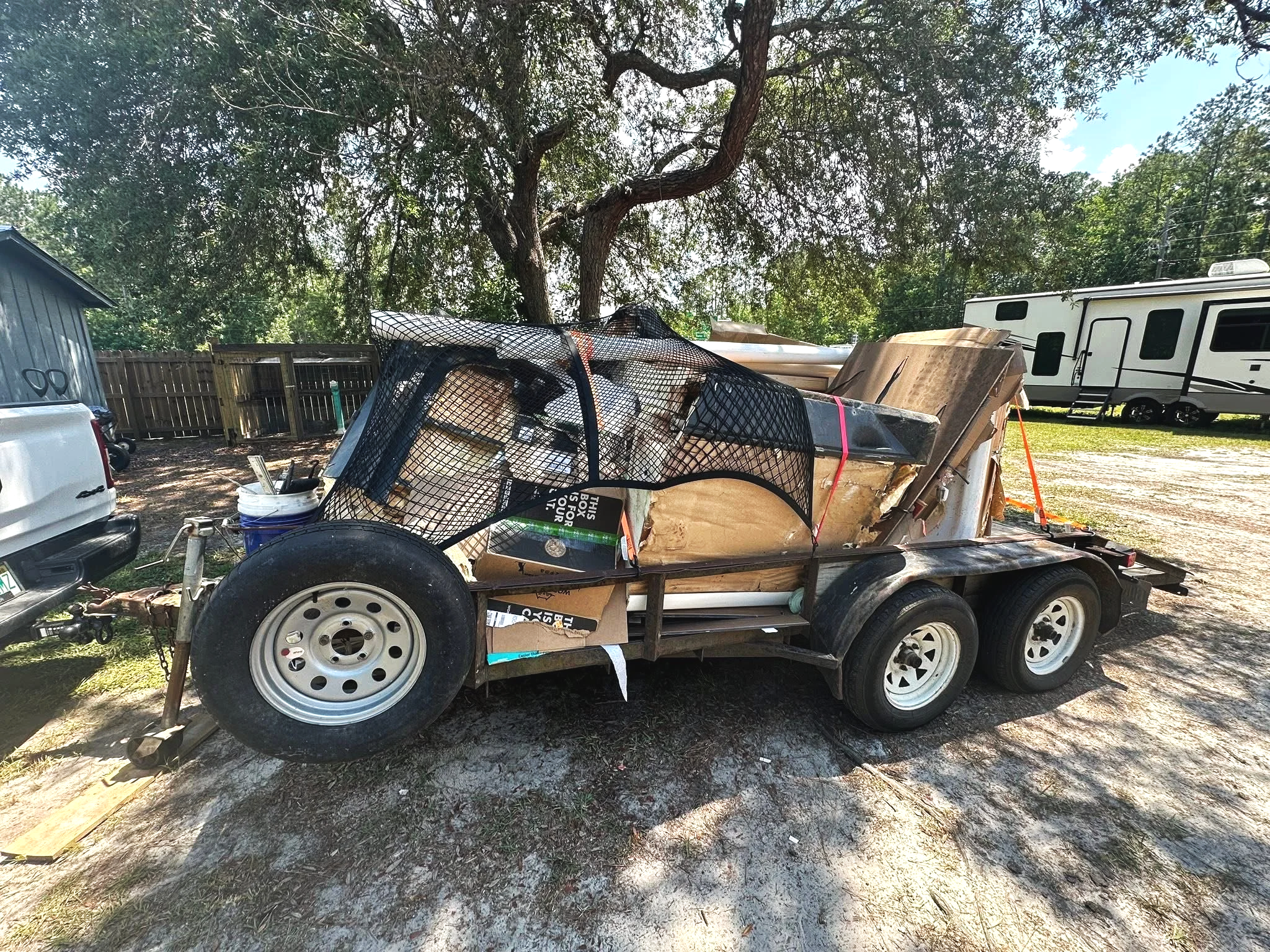 A trailer loaded with construction materials, tools, and cardboard boxes, parked outdoors near a tree and a white building with a fence and RVs in the background.
