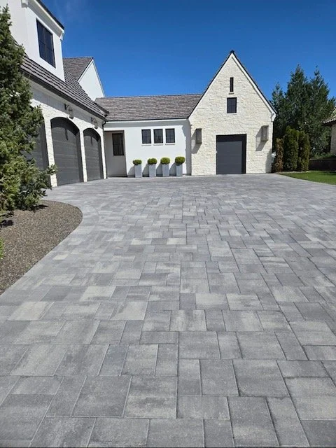 Modern house with white stone exterior and a large paved driveway leading to garage doors, surrounded by neatly landscaped yard under a clear blue sky.