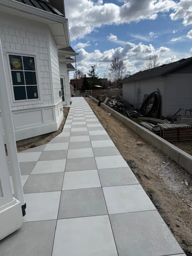 Newly constructed sidewalk made of large white and gray tiles running alongside a white house with a decorative bay window, with a partly cloudy sky overhead.