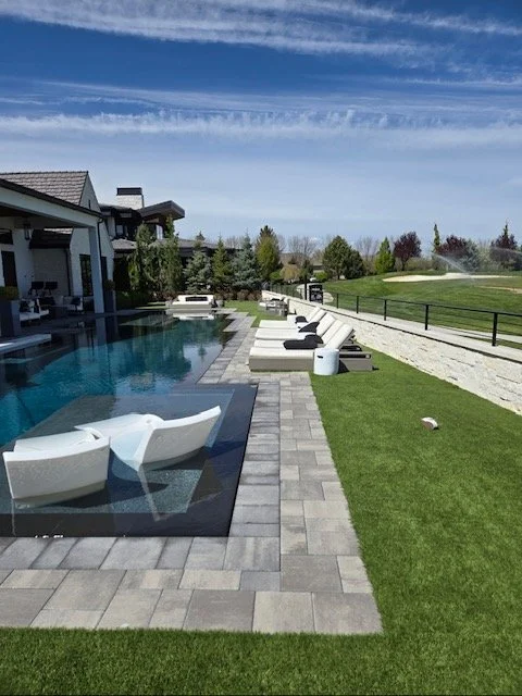 Luxury backyard pool area with white lounge chairs, a flat grassy lawn, and a landscaped garden under a clear blue sky.