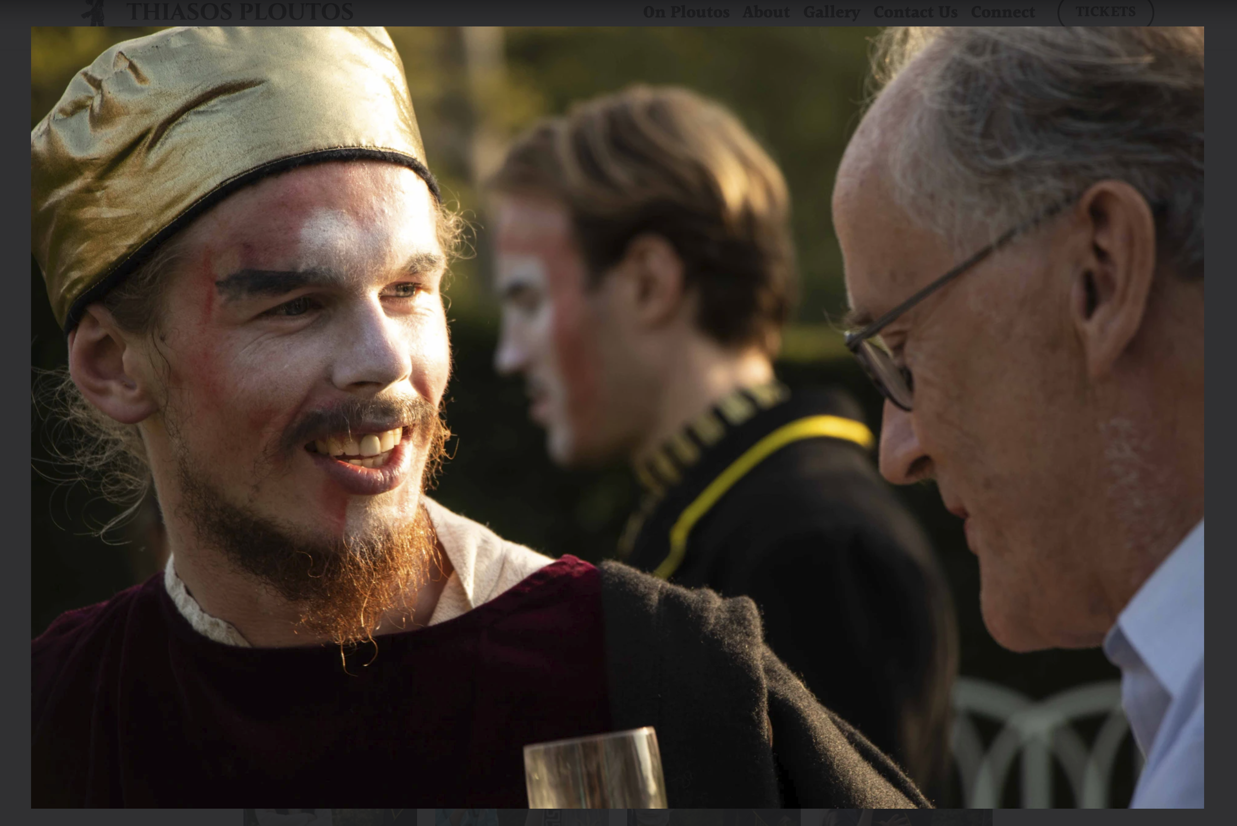 Three men at an outdoor gathering, two of them are engaged in conversation. One man has a gold hat, tan clothing, and a beard, smiling; the second man has glasses and gray hair, also smiling; the third man in the background has light brown hair and is dressed in a dark outfit with yellow accents.