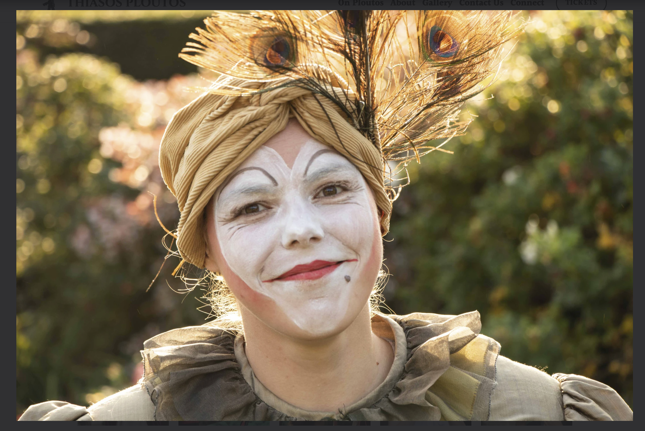 Person dressed as a clown or mime outdoors, wearing a beige headscarf with a peacock feather, white face paint with expressive eyebrows and red lipstick, smiling warmly against a blurred background of trees and foliage.