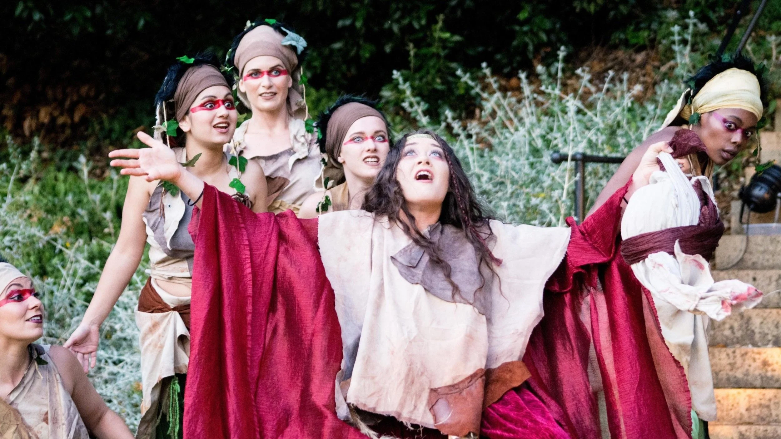 Actors on stage performing a theatrical scene dressed in costumes resembling forest or tribal attire with face paint, with greenery and stairs in the background.