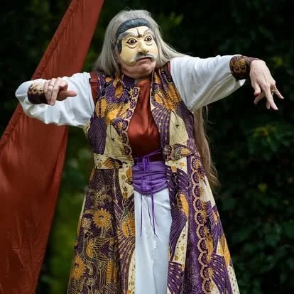 A person dressed in a theatrical costume with a mask that has exaggerated facial features, wearing traditional clothing with a patterned vest, long white sleeves, and a sash. The background shows a red curtain and green foliage.