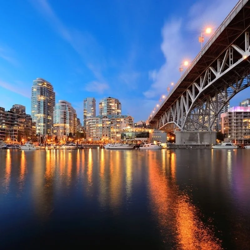 City skyline with modern high-rise buildings and a large bridge over water, illuminated during twilight. |  Moscone Developments - Custom Home Builder in Vancouver