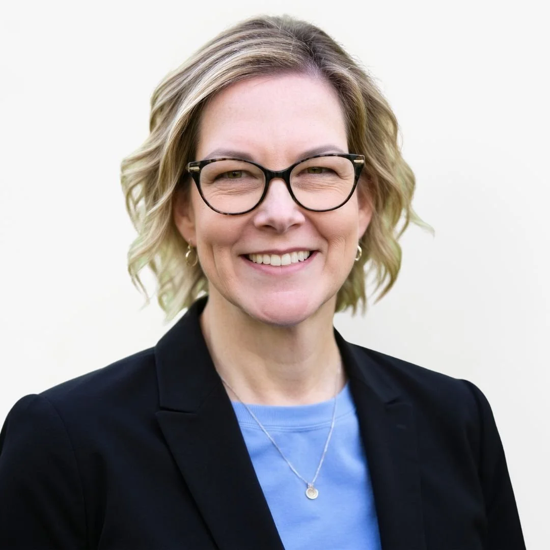 A woman with short hair, wearing glasses, gold jewelry, and a dark blazer, smiling at the camera against a plain white background.