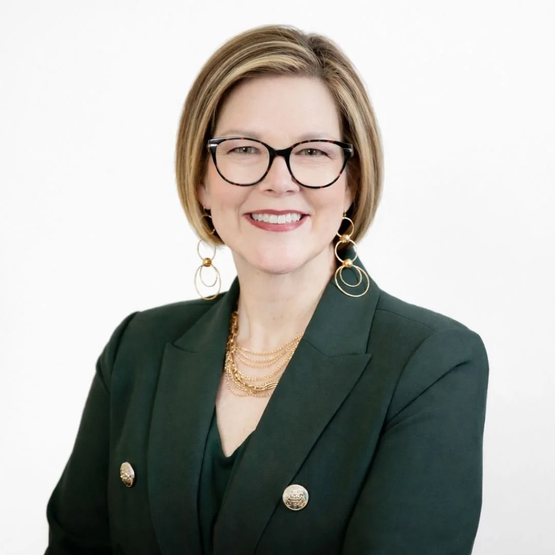 A woman with short hair, wearing glasses, gold jewelry, and a dark blazer, smiling at the camera against a plain white background.