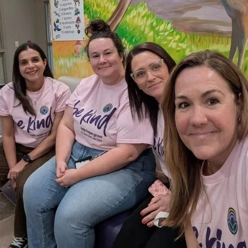 Four ladies sitting facing the camera smiling wearing "Be Kind" pink t-shirts