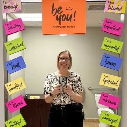 Woman standing in front of a mirror taking a selfie and around the border of the mirror are affirmations like "I am enough" and "I am strong"