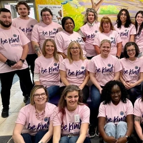 Group photo of LCC team members sitting together and smiling while wearing pink Be Kind t-shirts