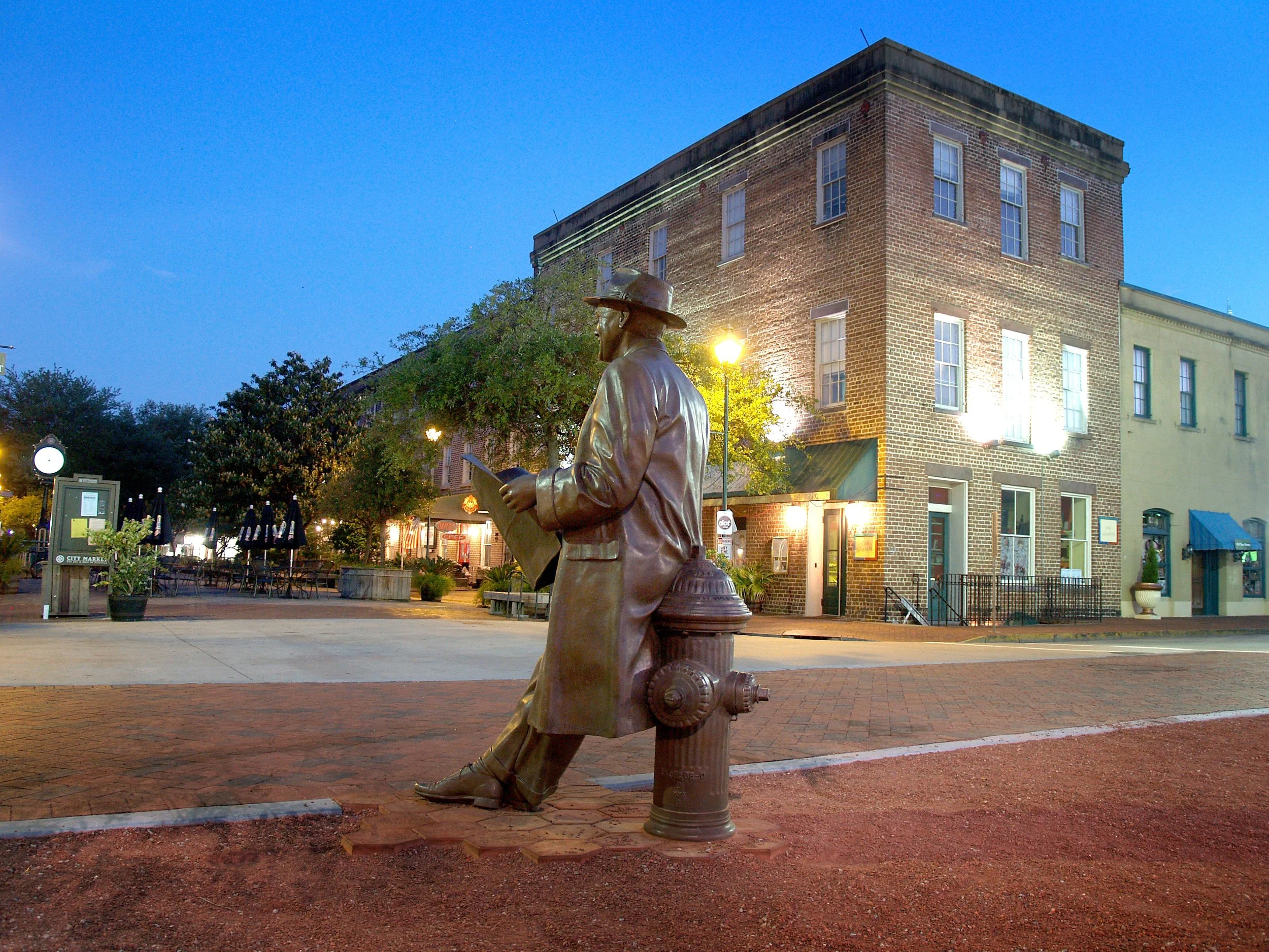 A statue of a man wearing a hat and overcoat sitting on a fire hydrant, reading a newspaper, in a small town or city square at dusk with brick buildings and streetlights.