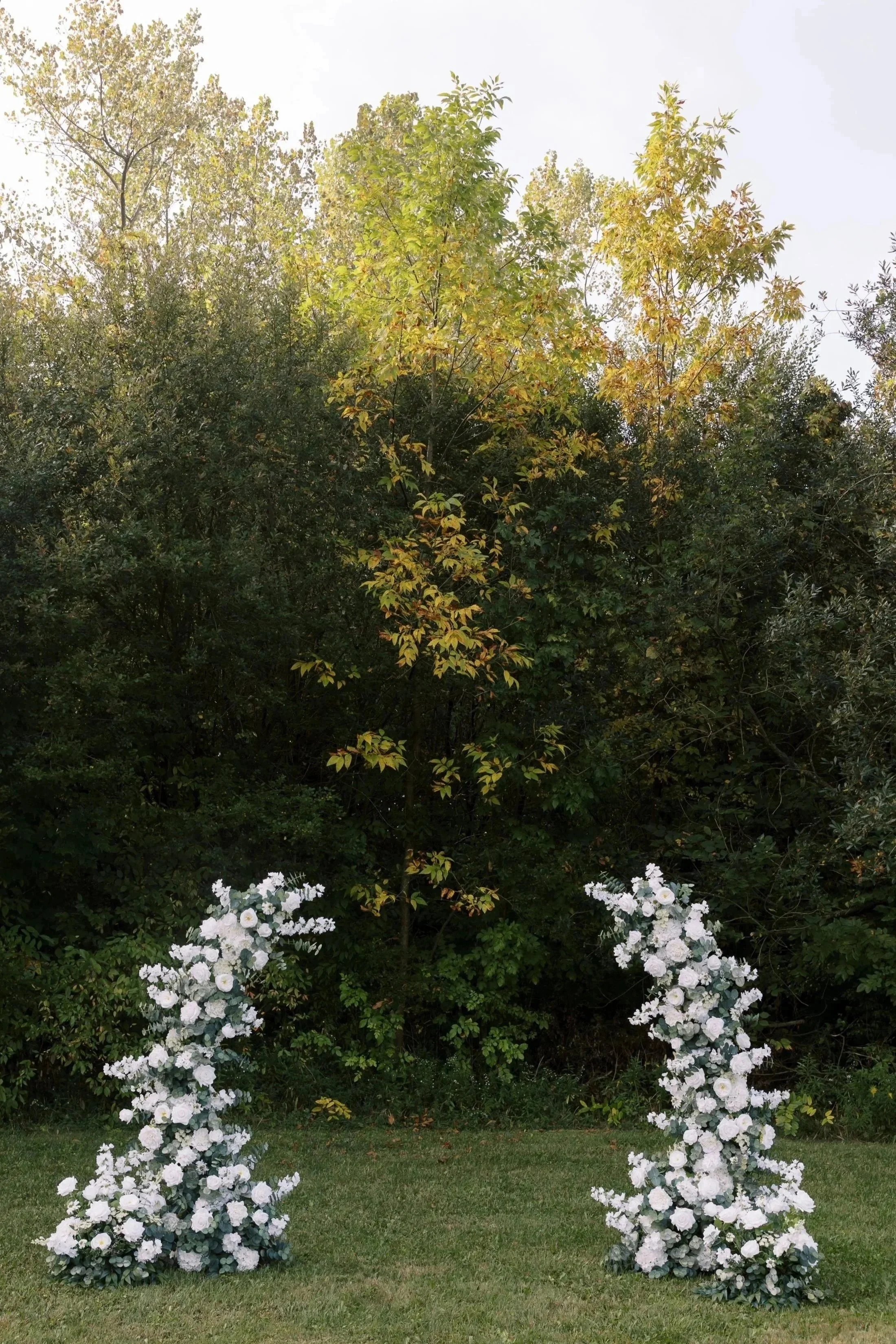 Two tall floral arrangements with white flowers arching on either side of a grassy lawn, with dense trees and bushes in the background.