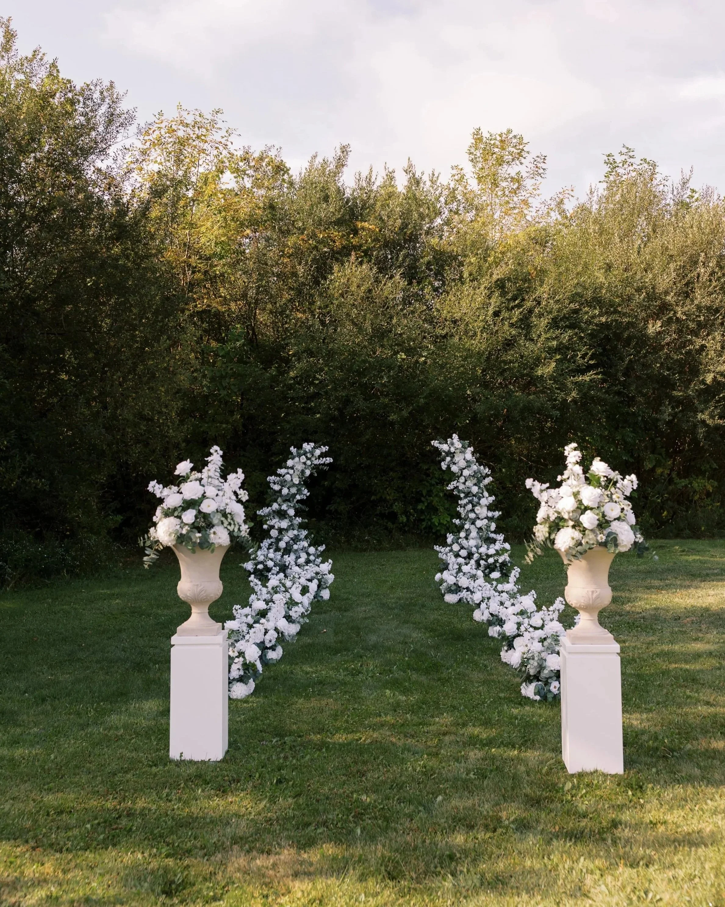 Two white floral arrangements in tall white vases and four floral arches decorate an outdoor lawn, with greenery trees in the background.