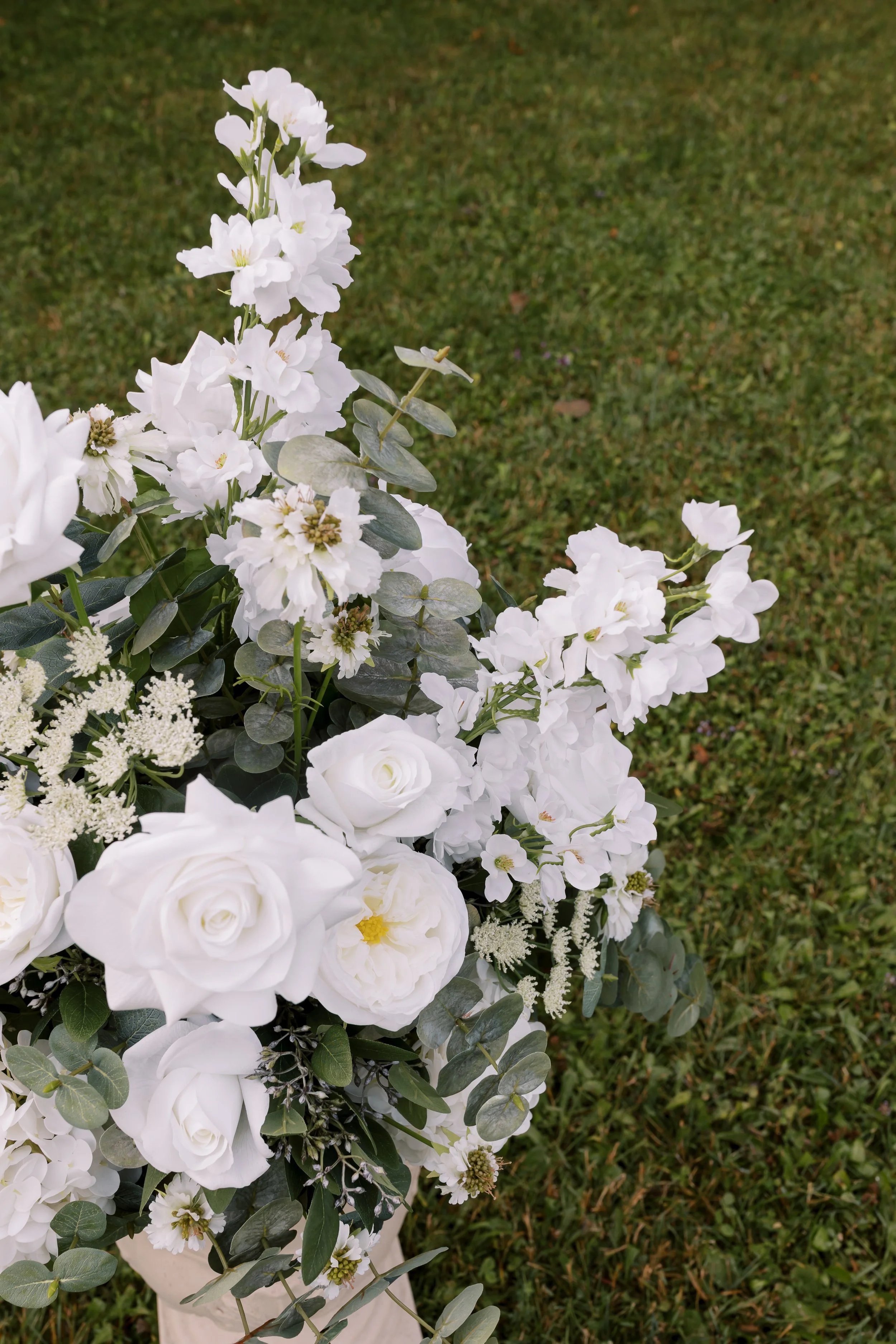 White floral arrangement with roses and other white flowers on a grassy background.