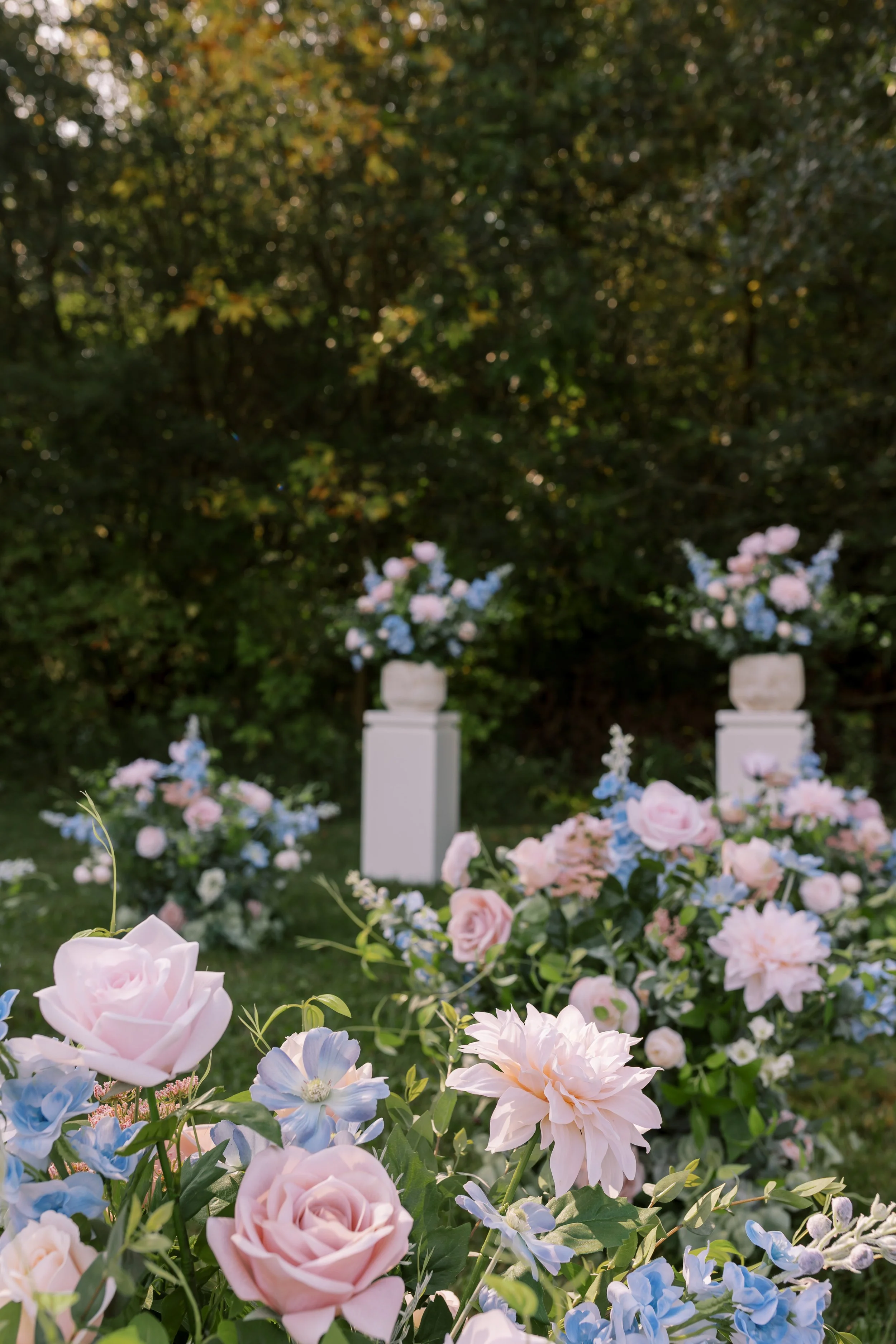 Floral arrangement with pink and blue flowers, possibly for a wedding or outdoor event, set against a backdrop of greenery and trees.