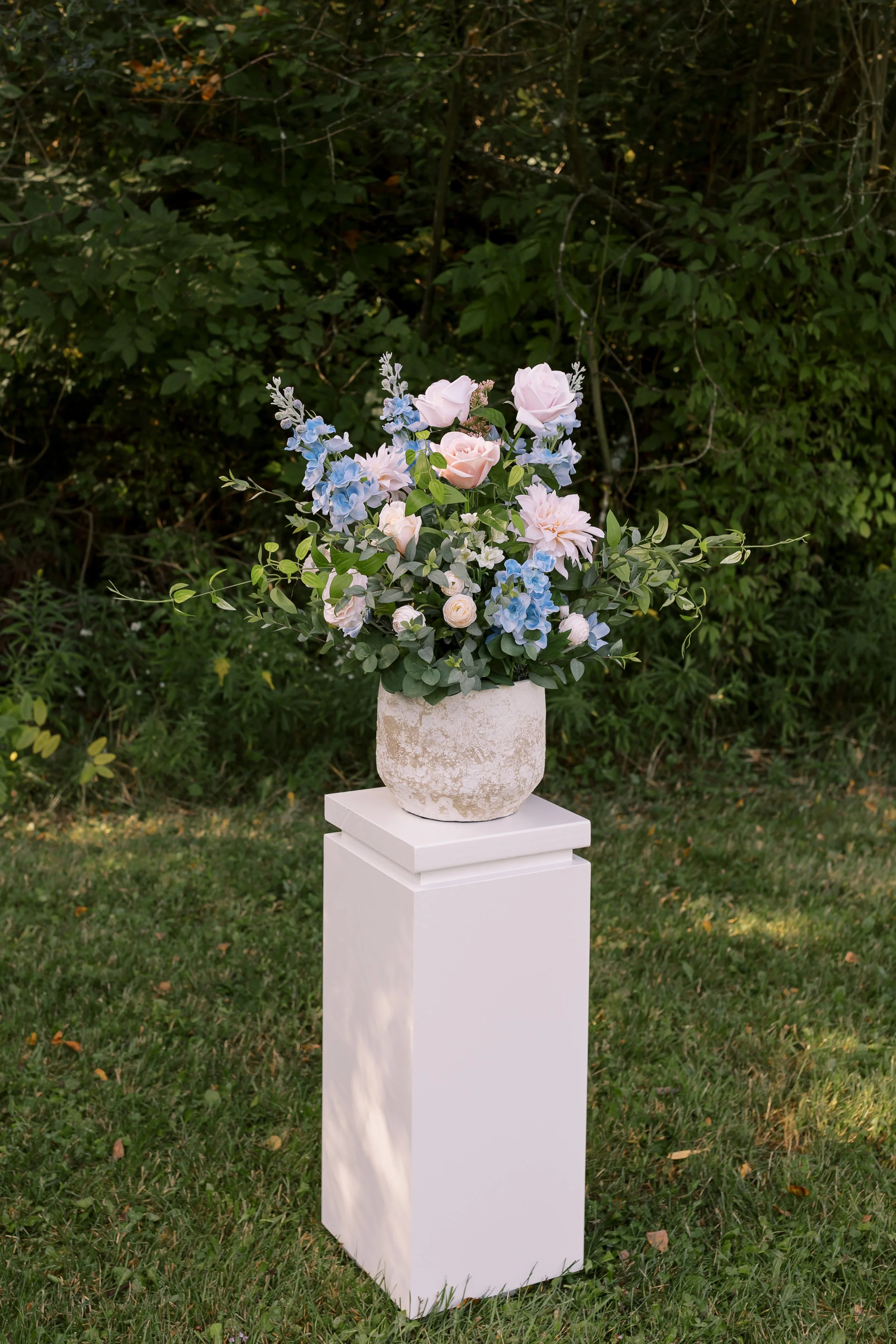 A floral arrangement of pink and blue flowers in a rustic stone pot, placed on a white pedestal outdoors with green bushes in the background.