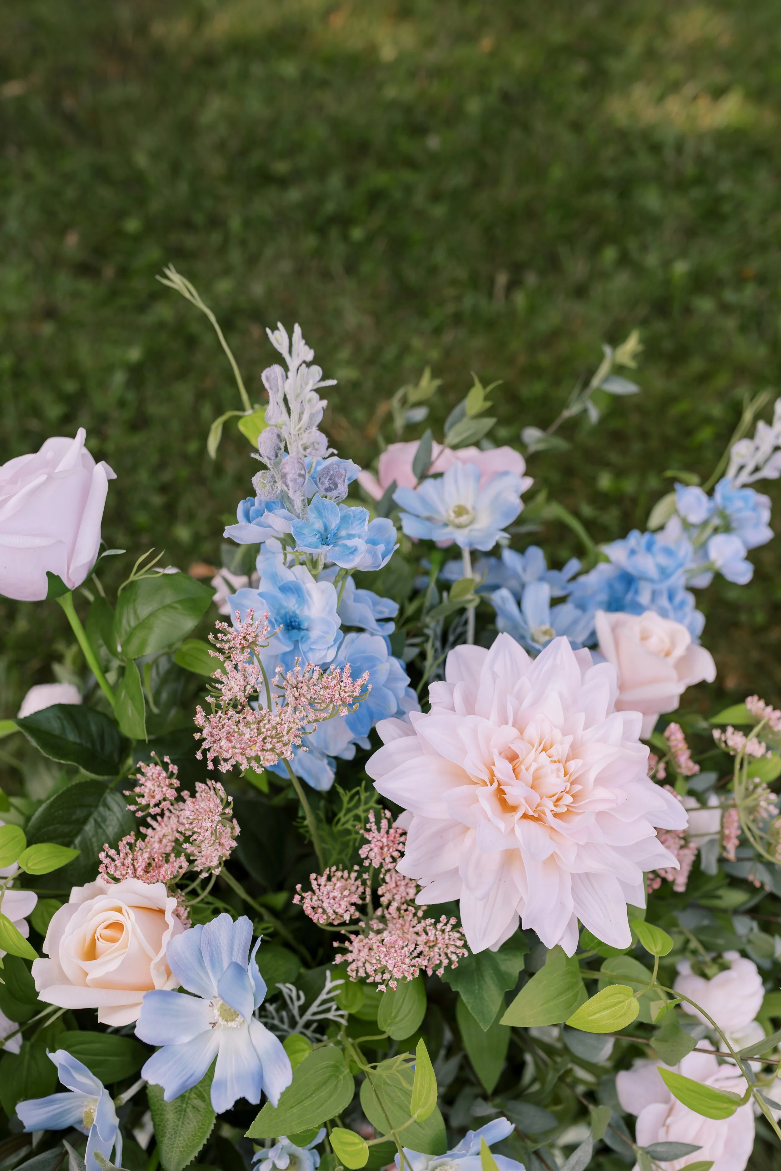 A bouquet of pastel pink, blue, and white flowers with green leaves on a grass background.