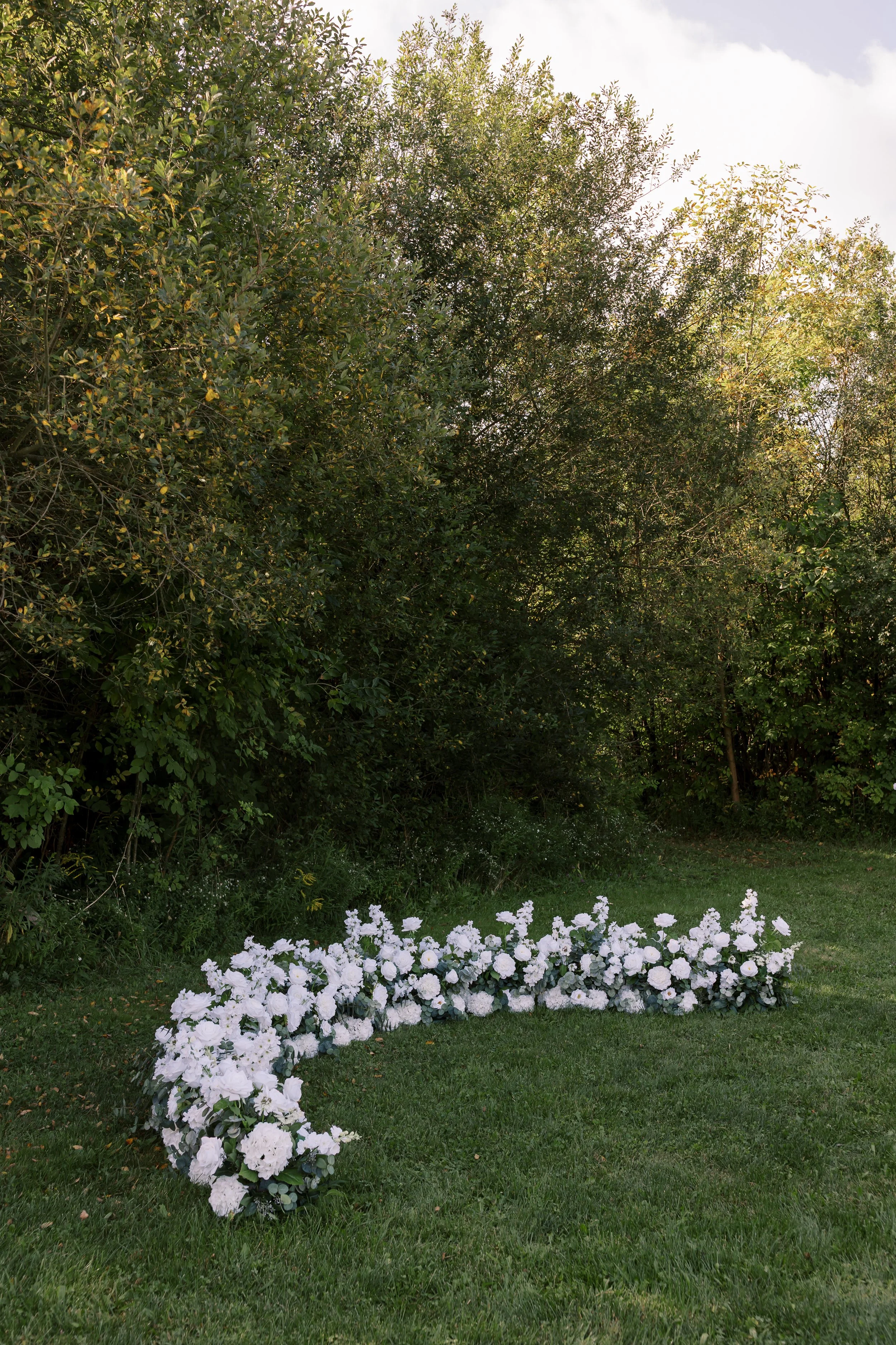 White floral arrangement on grass in front of dense green bushes and trees.