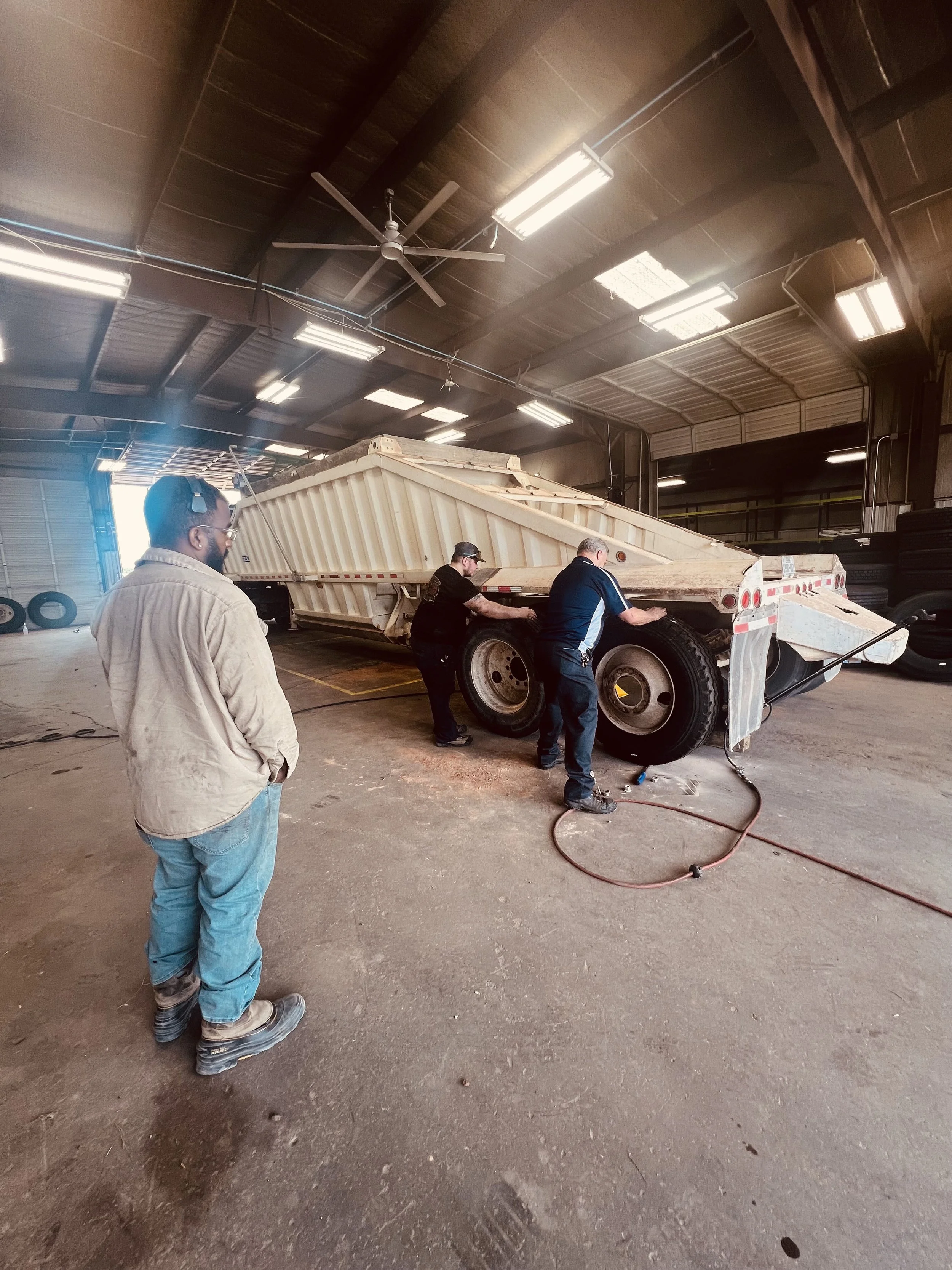 Three workers are inspecting or repairing a large dump truck inside a warehouse or garage.