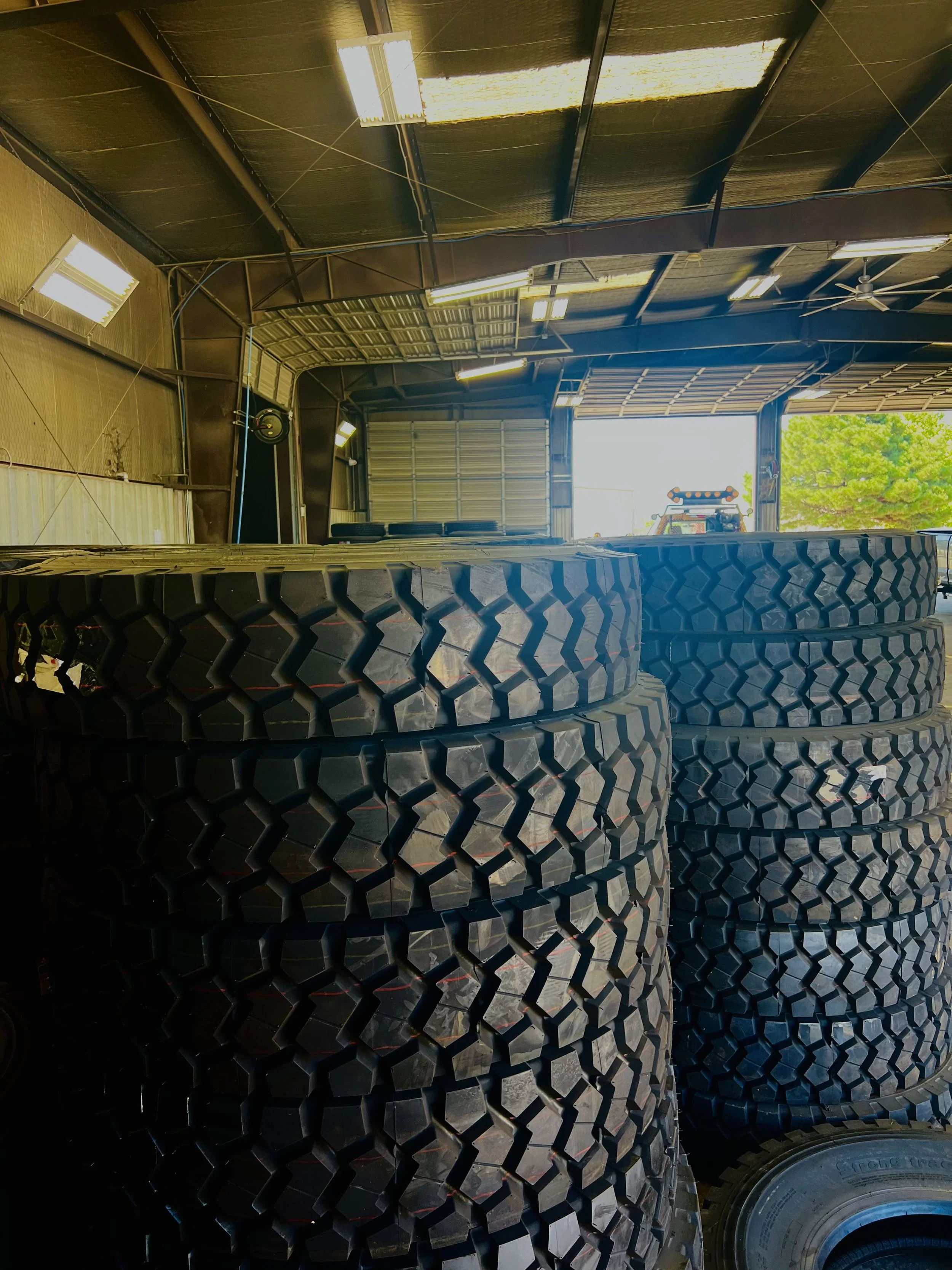 Stacked large off-road tires inside a warehouse with open doors, with a green tree and sky visible outside.