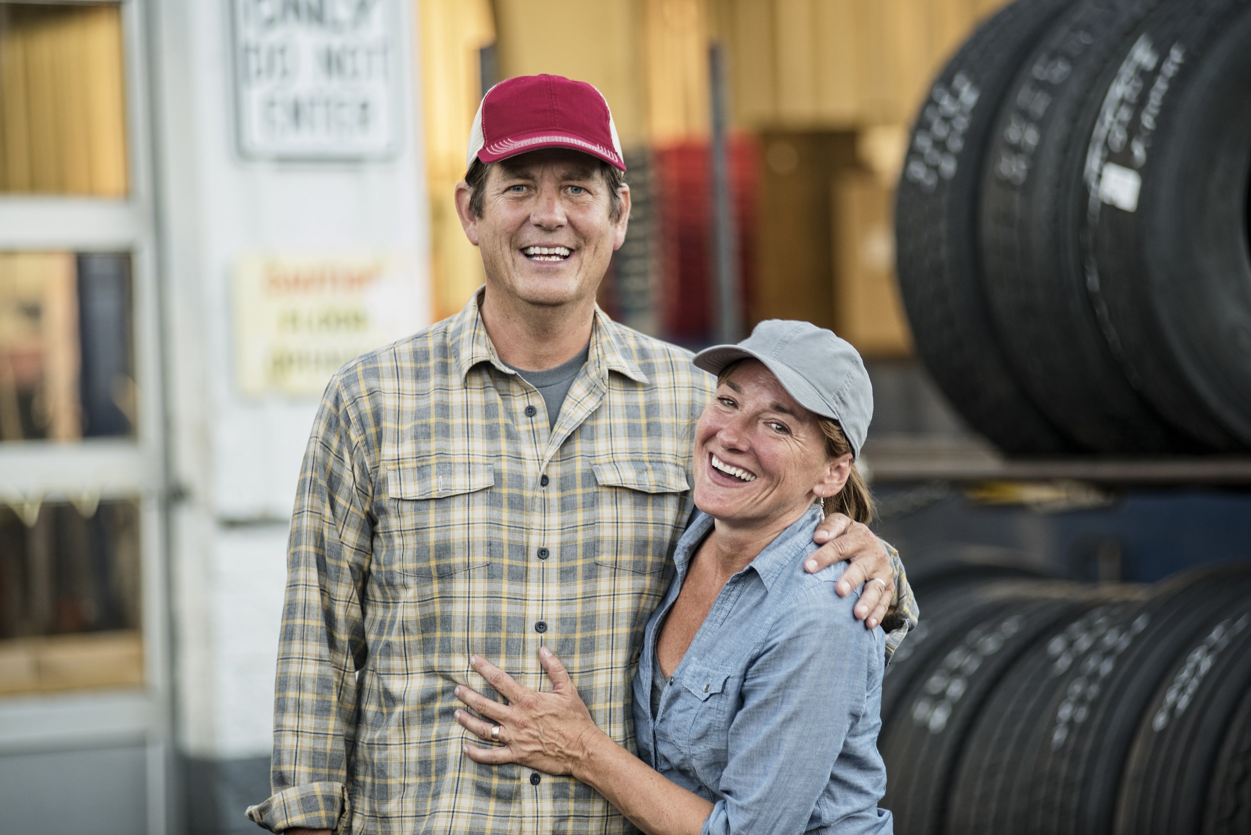 A smiling couple, a man and a woman, standing together in a tire shop or garage, with tires stacked on shelves behind them.