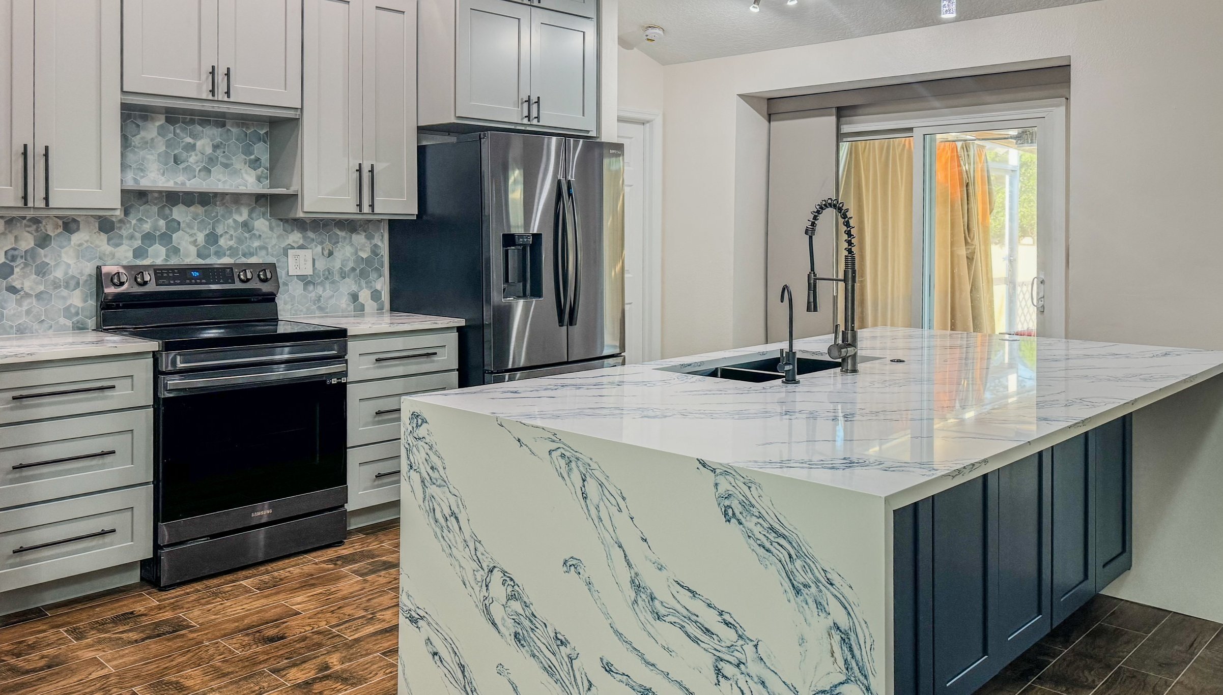 Modern kitchen with grey cabinets, a marble island with a black faucet, stainless steel refrigerator, and a stove against a patterned tile backsplash.