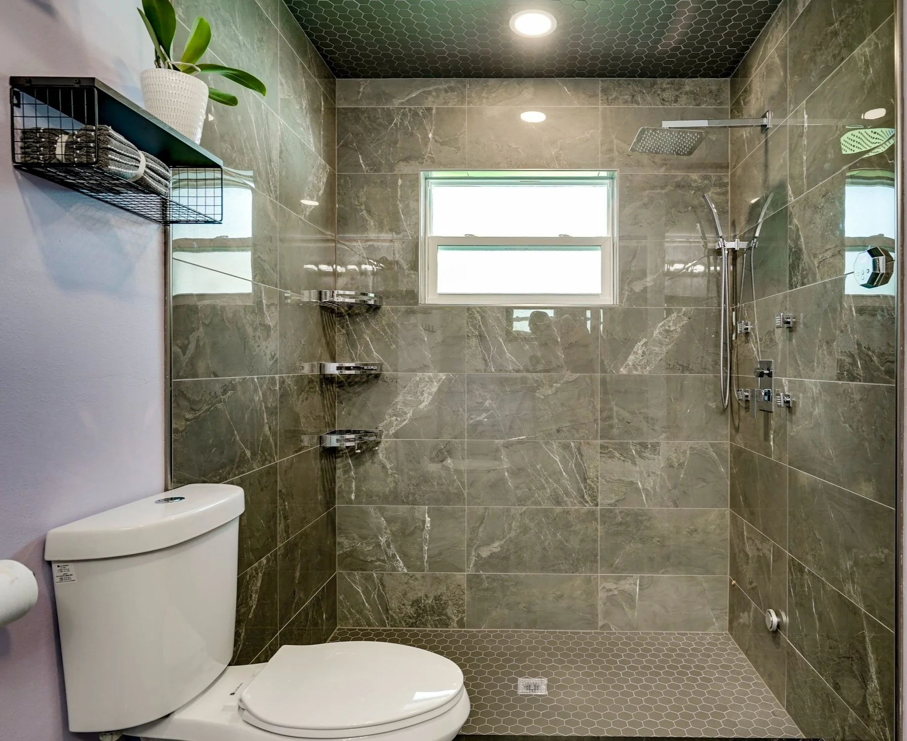 Bathroom with tiled walls and floor, a toilet, a window, and a walk-in shower with a rain showerhead, handheld shower, and shower controls.