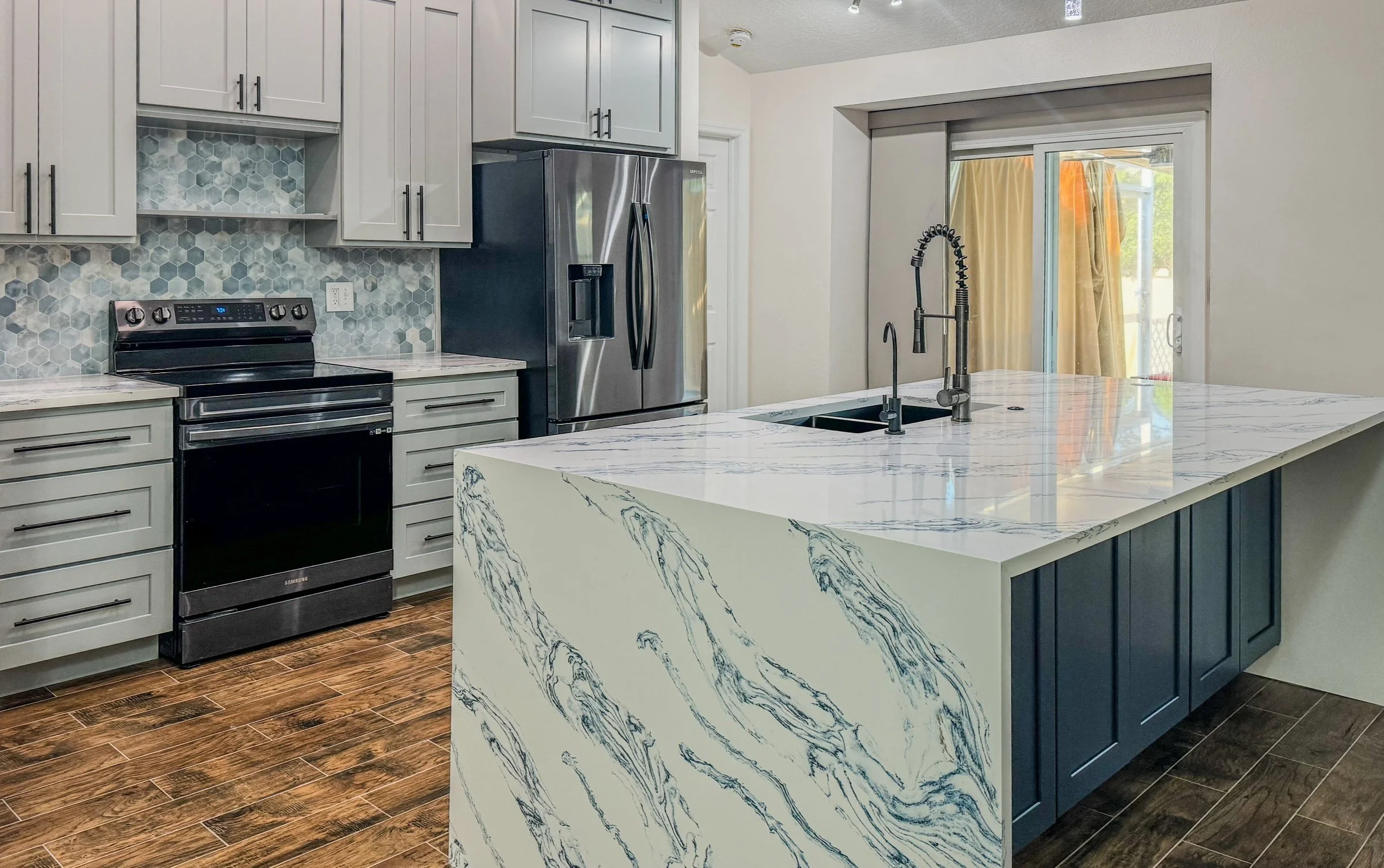Modern kitchen with white cabinetry, stainless steel appliances, a hexagonal tile backsplash, and a marble-patterned island with a black faucet. Sliding glass door leads outside.