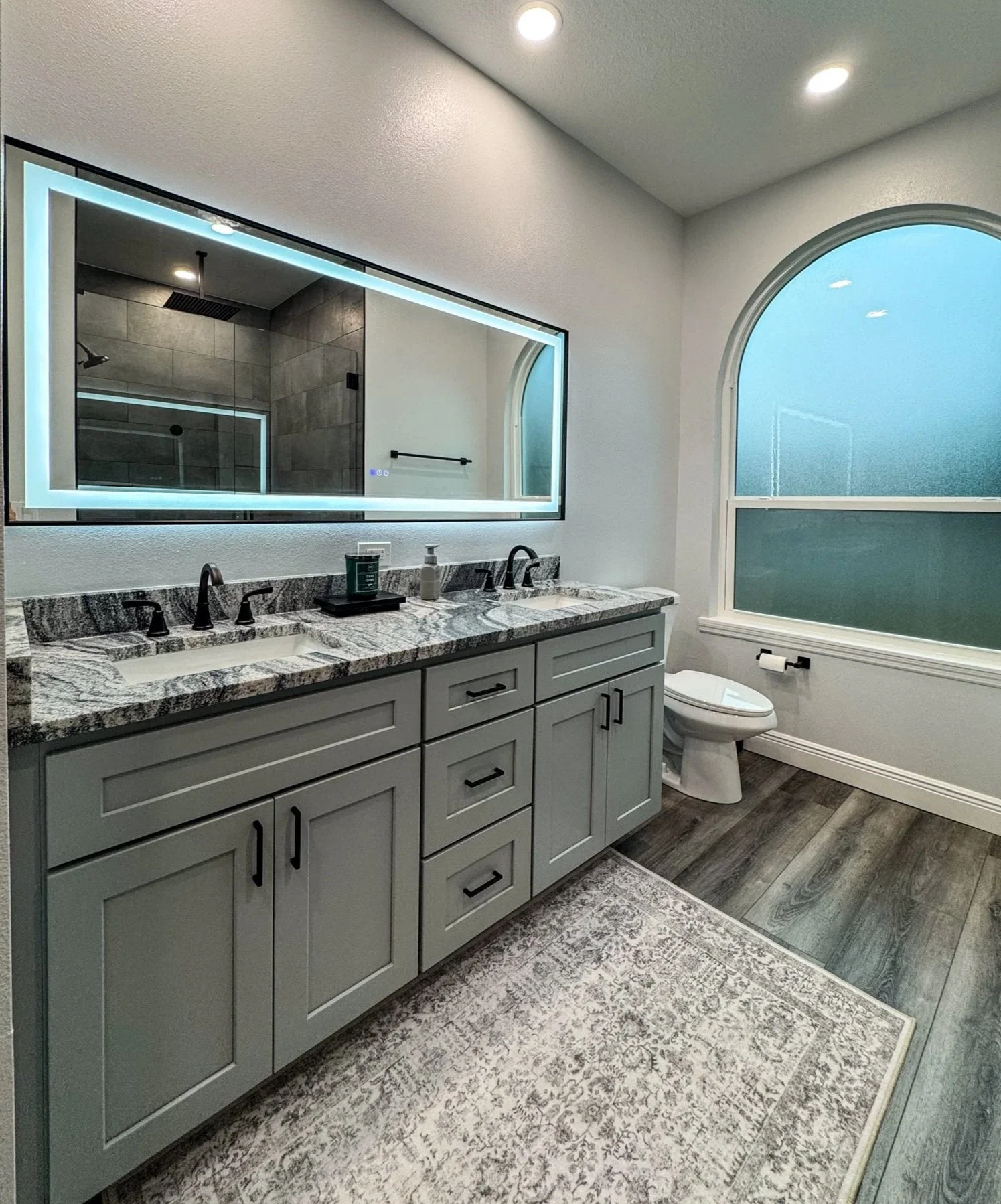 A modern bathroom with a gray vanity, granite countertop, and black fixtures. A large illuminated mirror above the sink, a window with frosted glass, and a partial view of a shower with gray tiles.