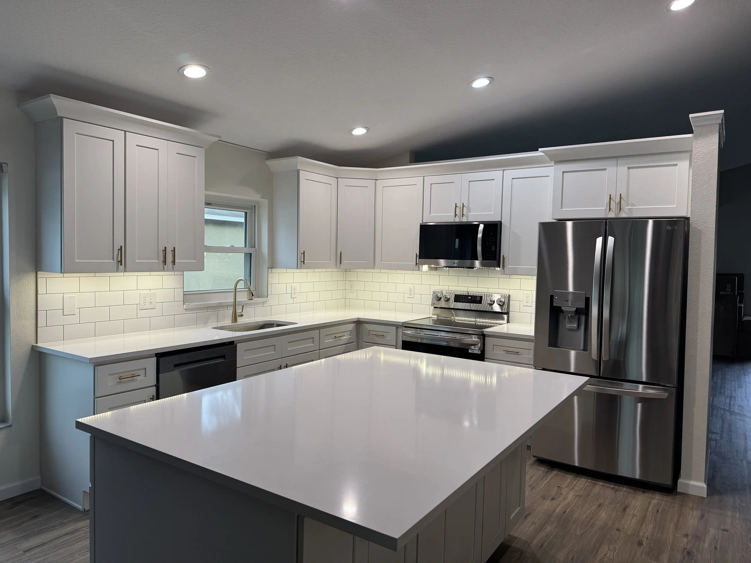 Modern kitchen with white cabinets, a white countertop island, stainless steel appliances including a refrigerator and microwave, a window above the sink, and recessed ceiling lights.