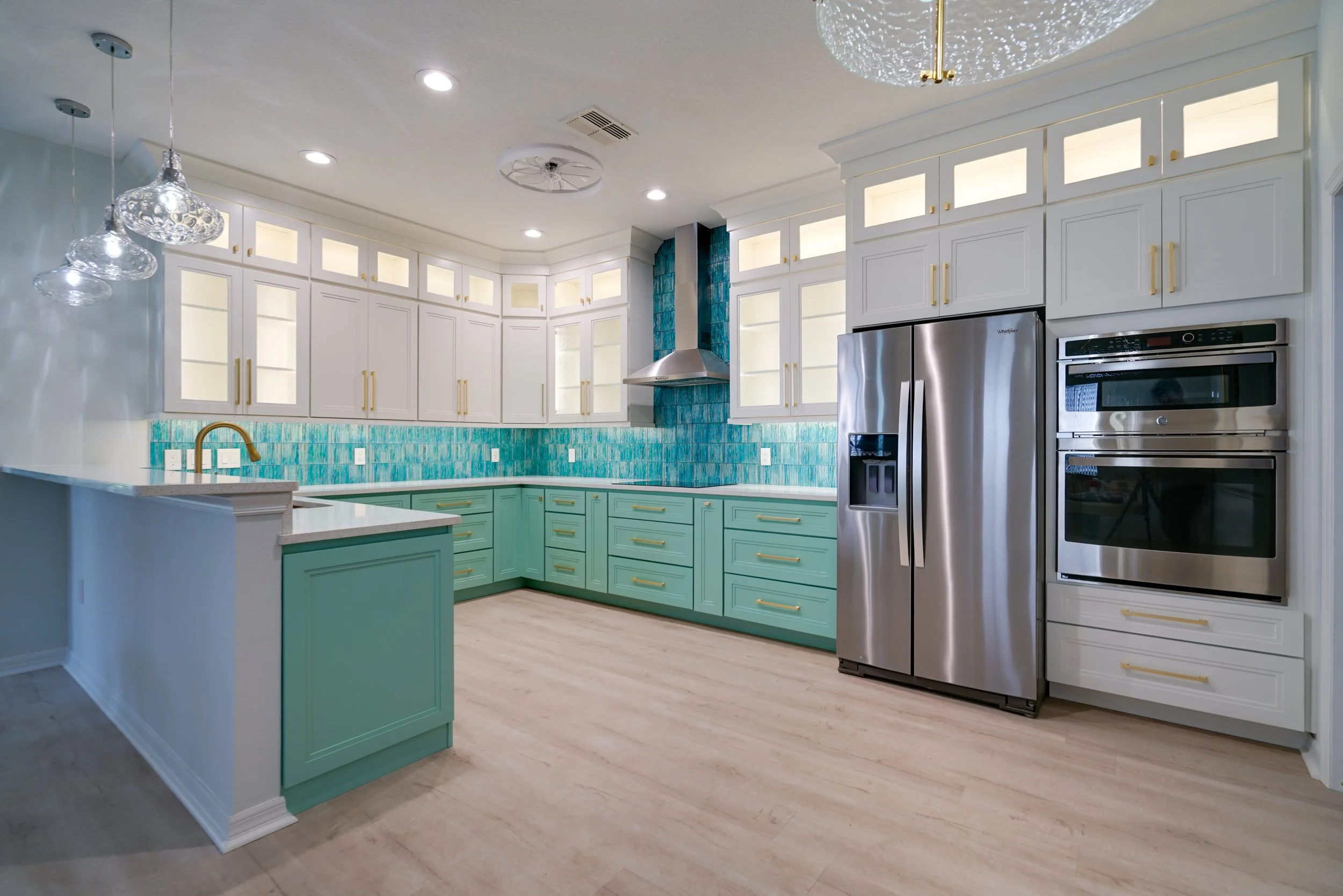 Modern kitchen with turquoise lower cabinets and white upper cabinets, stainless steel refrigerator and oven, turquoise tile backsplash, white countertop with gold fixtures, and three glass pendant lights.