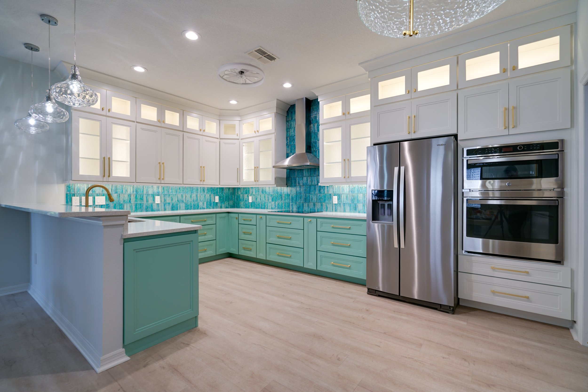 Modern kitchen with white upper cabinets, turquoise lower cabinets, stainless steel refrigerator, double wall ovens, blue tile backsplash, and a light-colored wood floor.