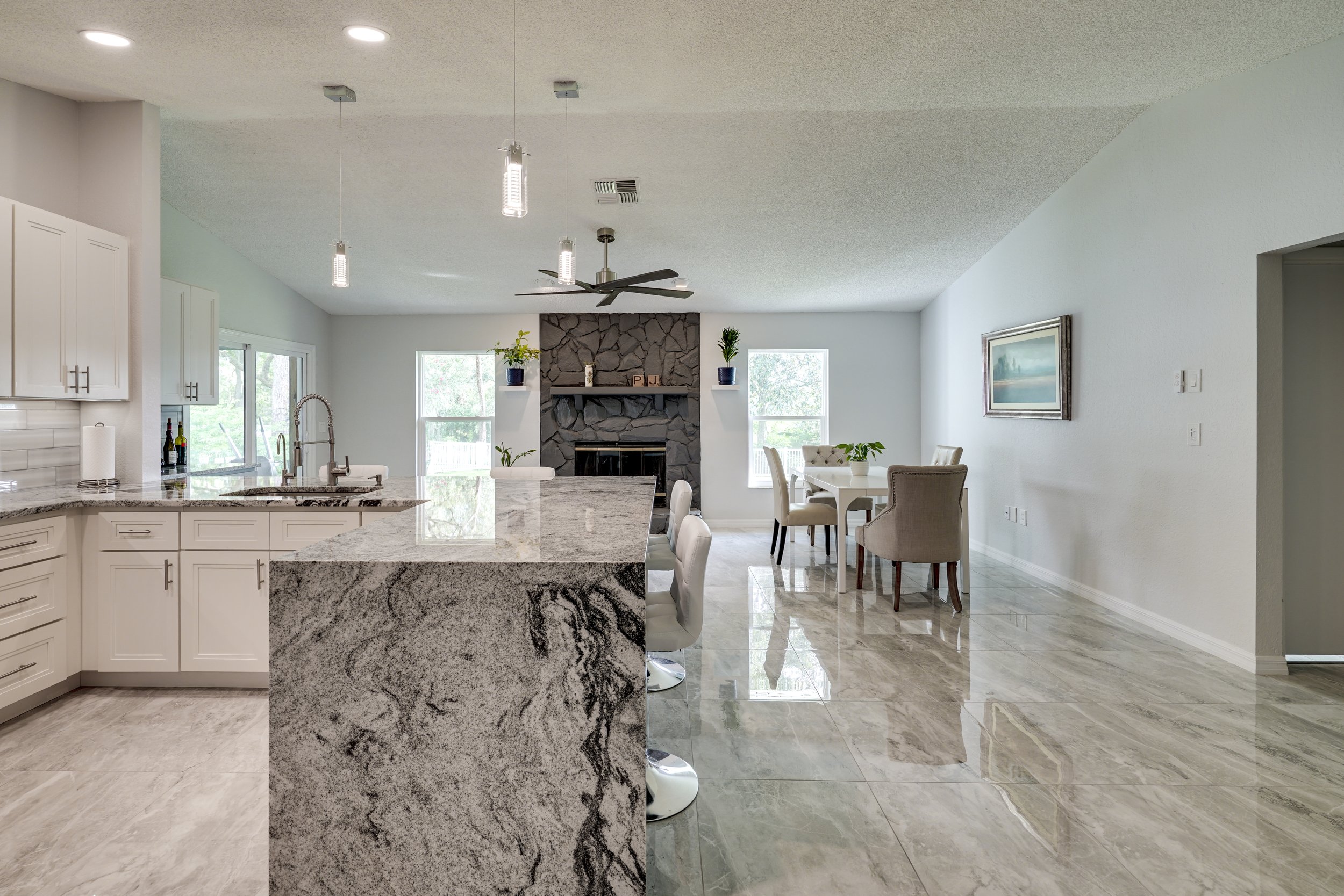 Open-concept kitchen and living room with granite countertops, white cabinets, a stone fireplace, dining area, and tiled flooring.