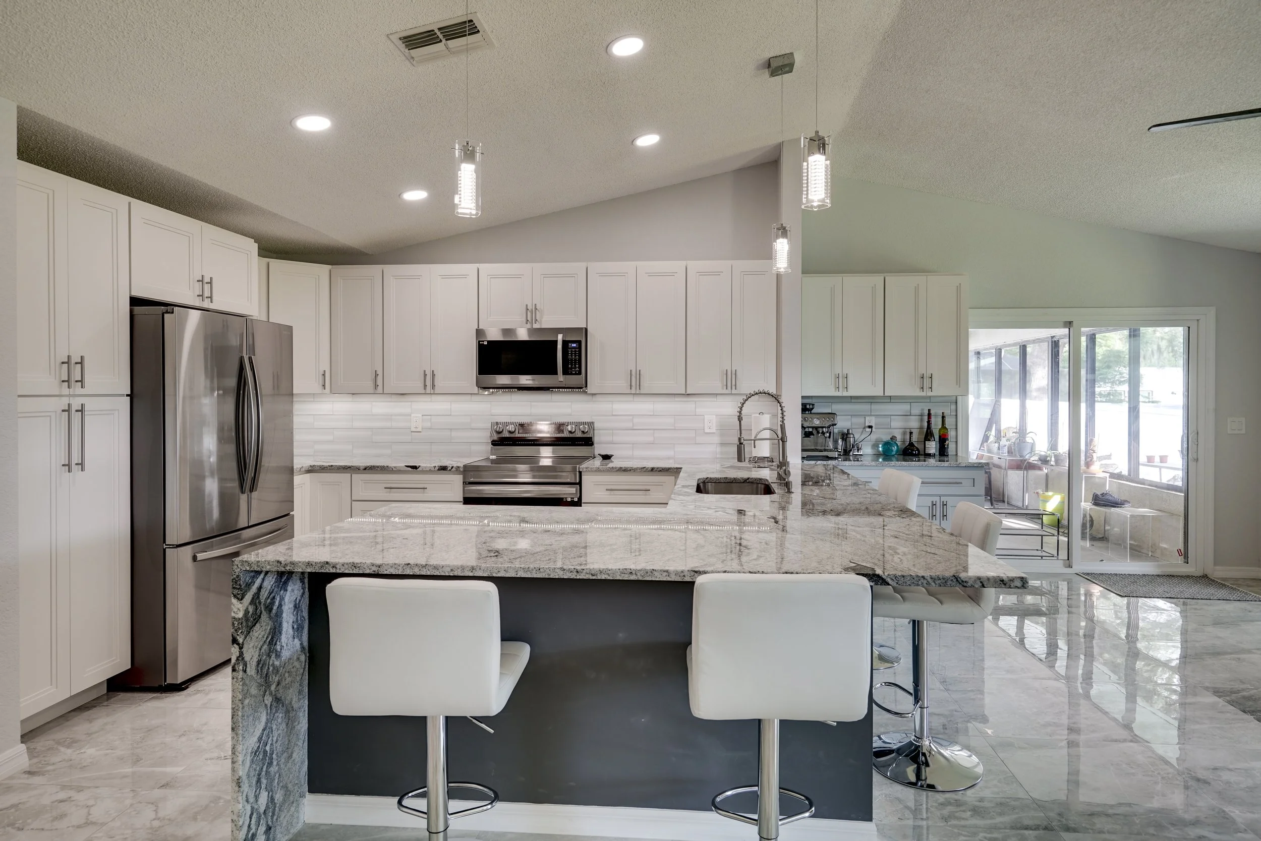 Modern kitchen with white cabinets, stainless steel appliances, granite countertops, and a central island with white bar stools. Recessed lighting and pendant lights hanging above the island. Sliding glass door leading to an outdoor area.