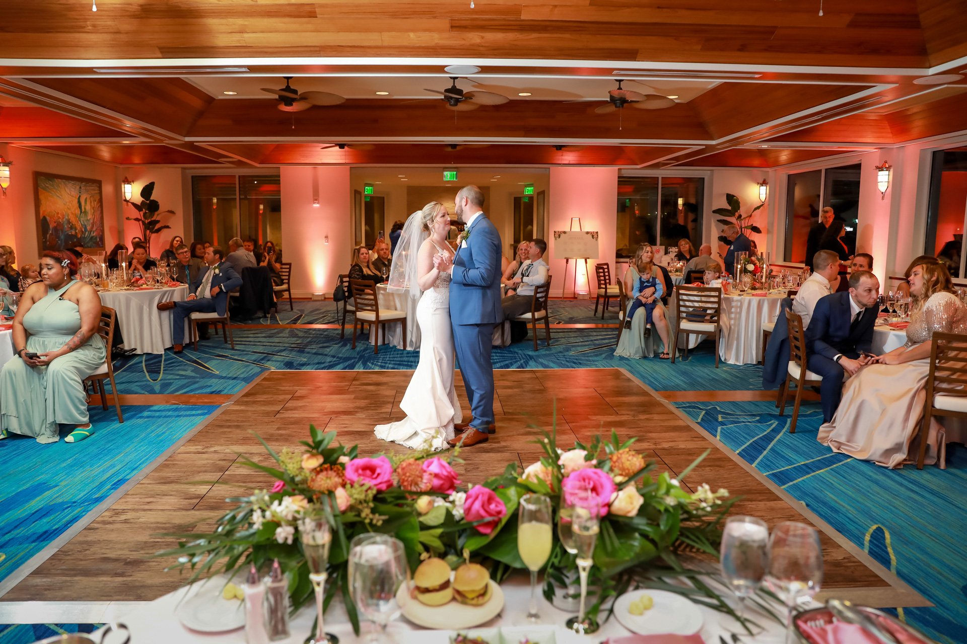 Bride and groom dancing at a wedding reception, surrounded by seated guests, with floral arrangements and dinner settings visible on tables.