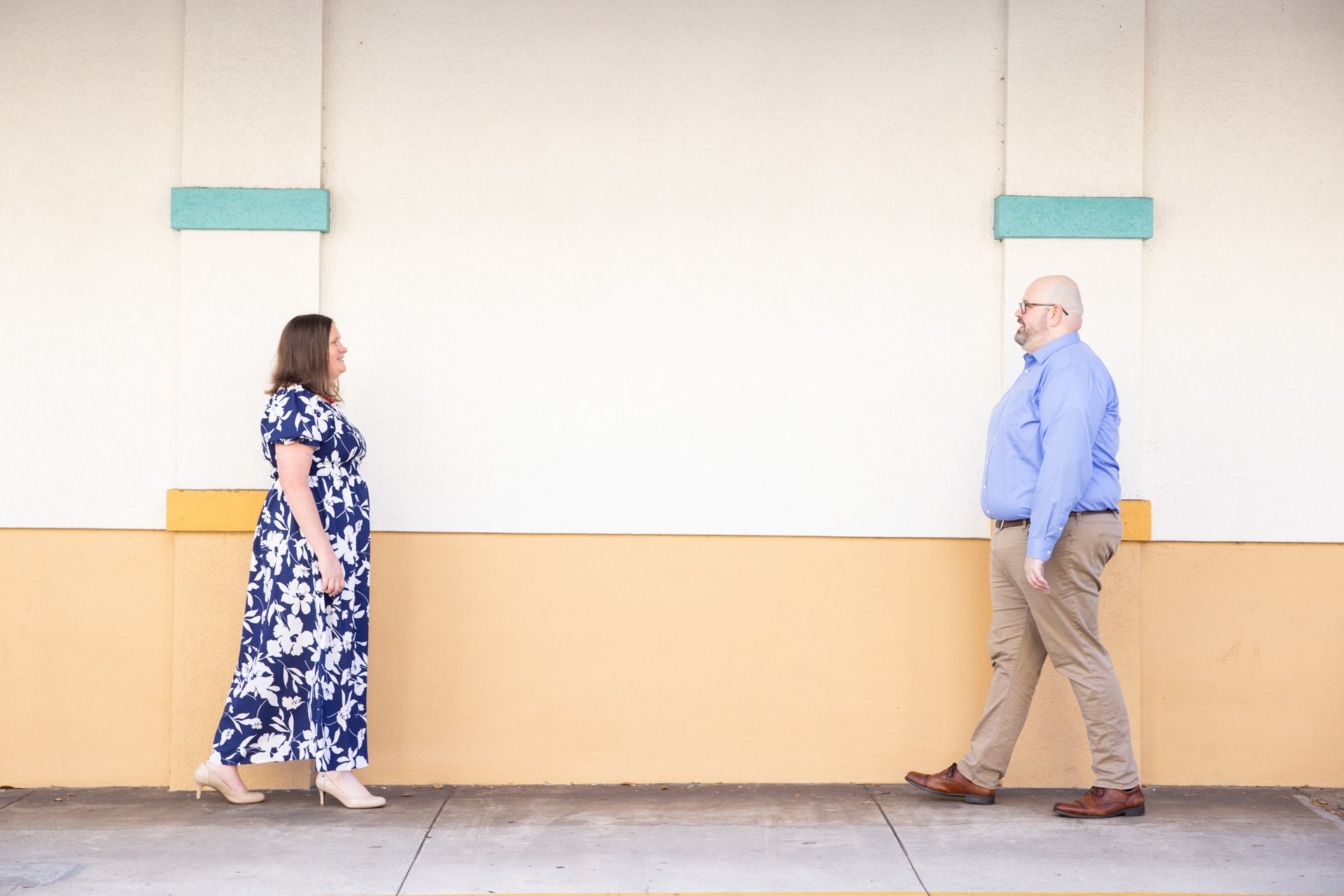 Two people walking towards each other on a sidewalk, with a beige and white wall in the background. The woman is wearing a blue floral dress and the man is wearing a blue shirt and beige pants.