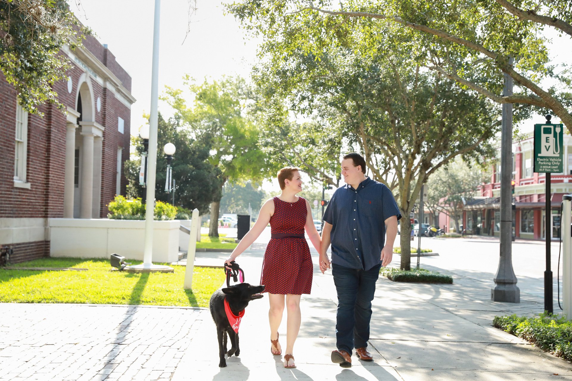 A couple holding hands walking with a black dog on a leash down a sunny street lined with trees and buildings, near an electric vehicle charging station.