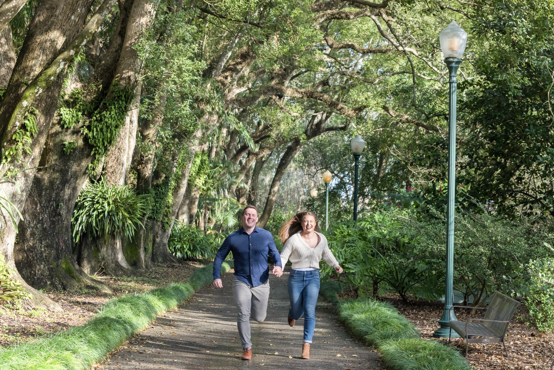 A couple holding hands and running joyfully on a tree-lined path with streetlamps and benches in a park.